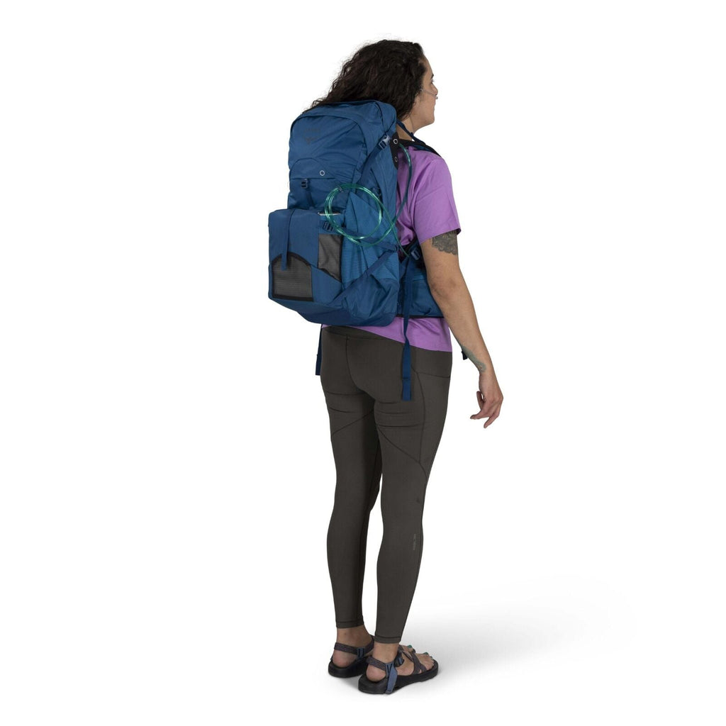 A person with long hair stands sideways, wearing a purple shirt, dark leggings, and sandals, carrying a large blue backpack. The tone is casual and outdoorsy.