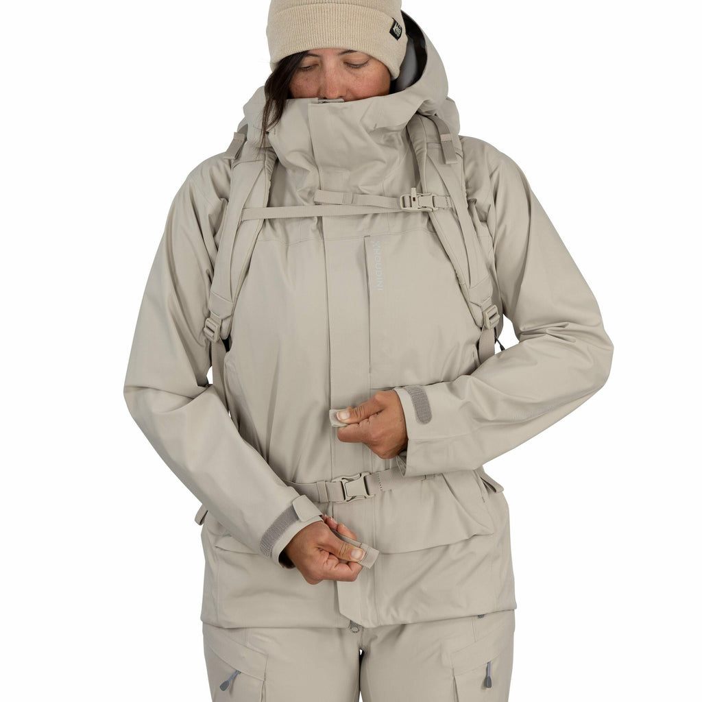 The image shows a person wearing a beige-colored outdoor outfit, which includes a jacket and matching pants. The jacket has multiple features such as a hood, adjustable straps, and several pockets, indicating it's designed for outdoor activities, possibly