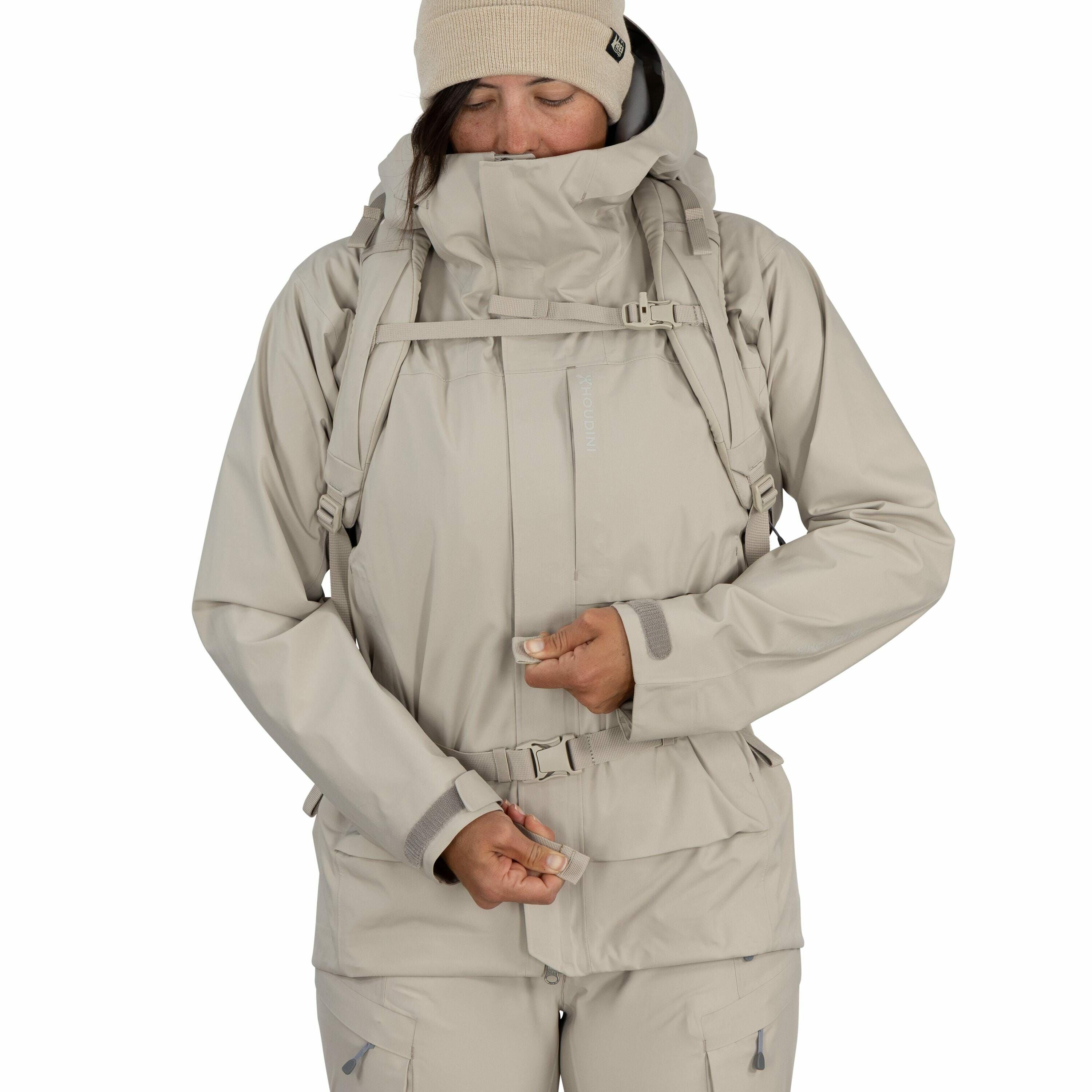 The image shows a person wearing a beige-colored outdoor outfit, which includes a jacket and matching pants. The jacket has multiple features such as a hood, adjustable straps, and several pockets, indicating it's designed for outdoor activities, possibly