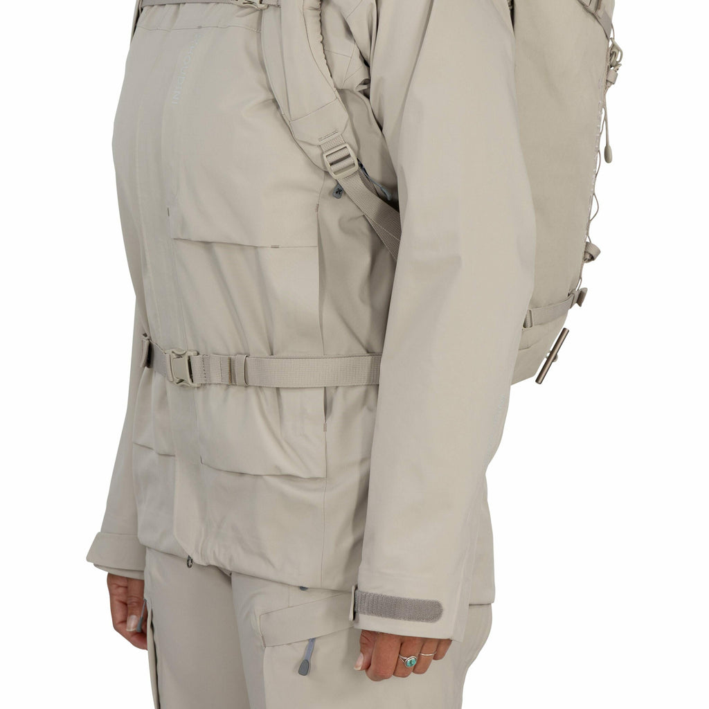 The image shows a person wearing a light beige outdoor jacket and matching pants, equipped with a backpack. The jacket has a front zipper closure, two large front pockets, and an adjustable waist belt for a better fit. The sleeves feature Velcro straps, a