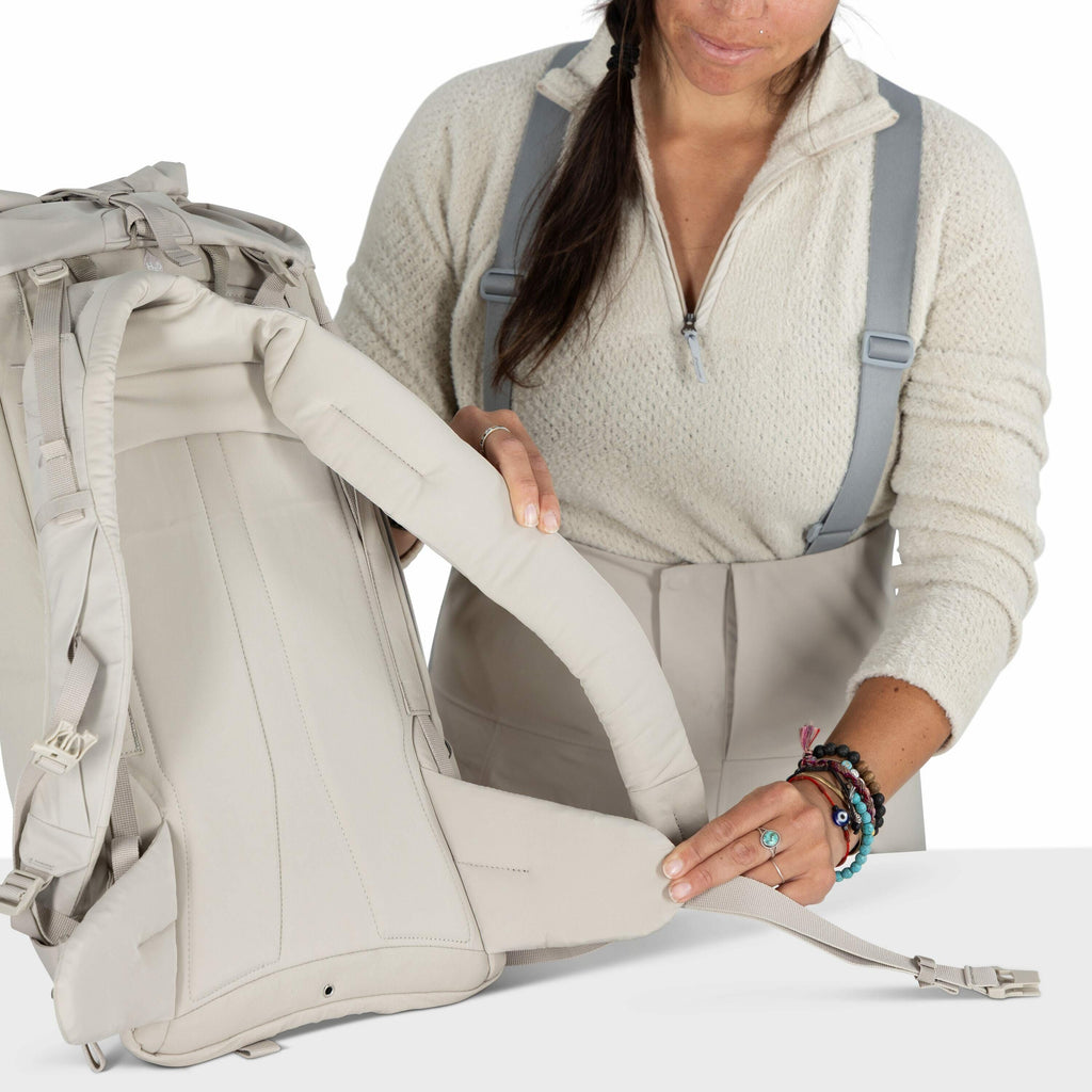 The image shows a woman demonstrating how to adjust the straps of a light-colored backpack. She is wearing a cream-colored sweater and gray suspenders, and her hair is loosely tied back. The focus is on her hands, which are manipulating the backpack's hip