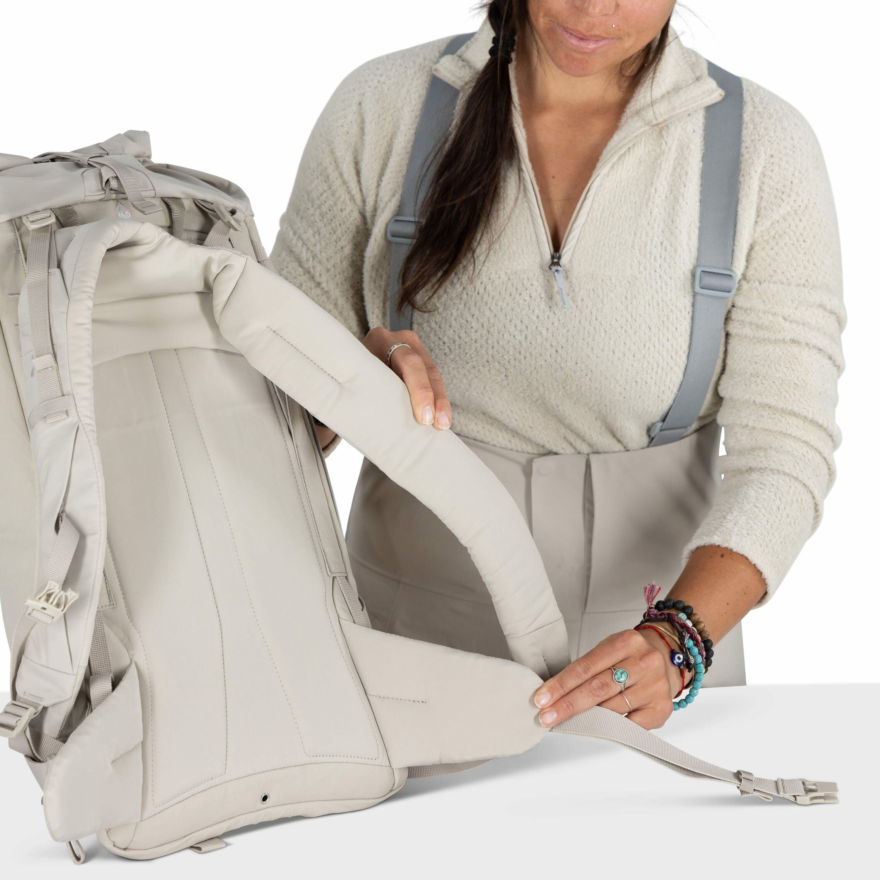 The image shows a woman demonstrating how to adjust the straps of a light-colored backpack. She is wearing a cream-colored sweater and gray suspenders, and her hair is loosely tied back. The focus is on her hands, which are manipulating the backpack's hip