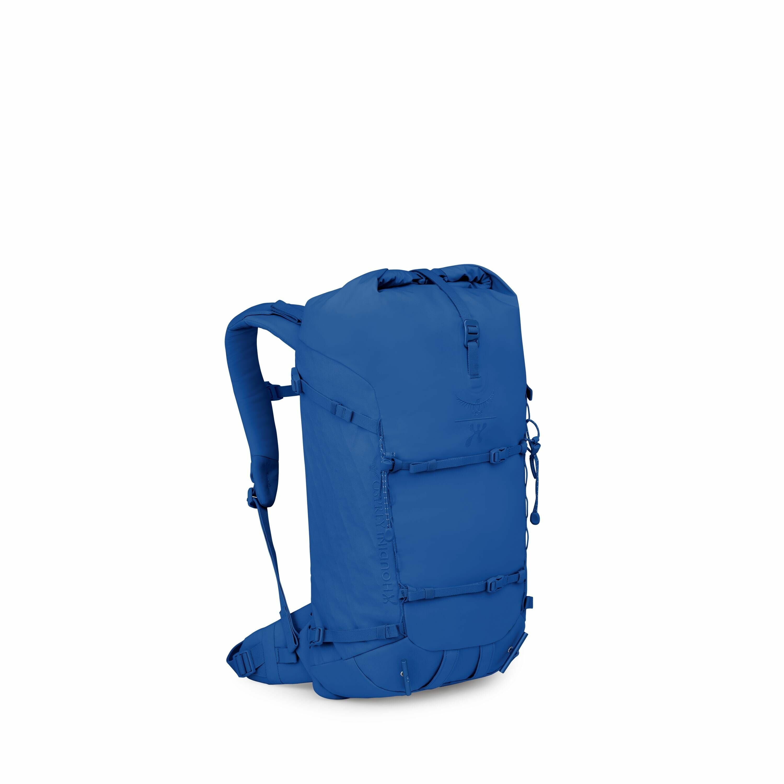 The image shows a blue backpack with a minimalist and sleek design. The backpack has a streamlined shape with a top-loading main compartment that appears to have a roll-top closure, as well as adjustable shoulder straps. There is a logo visible near the t