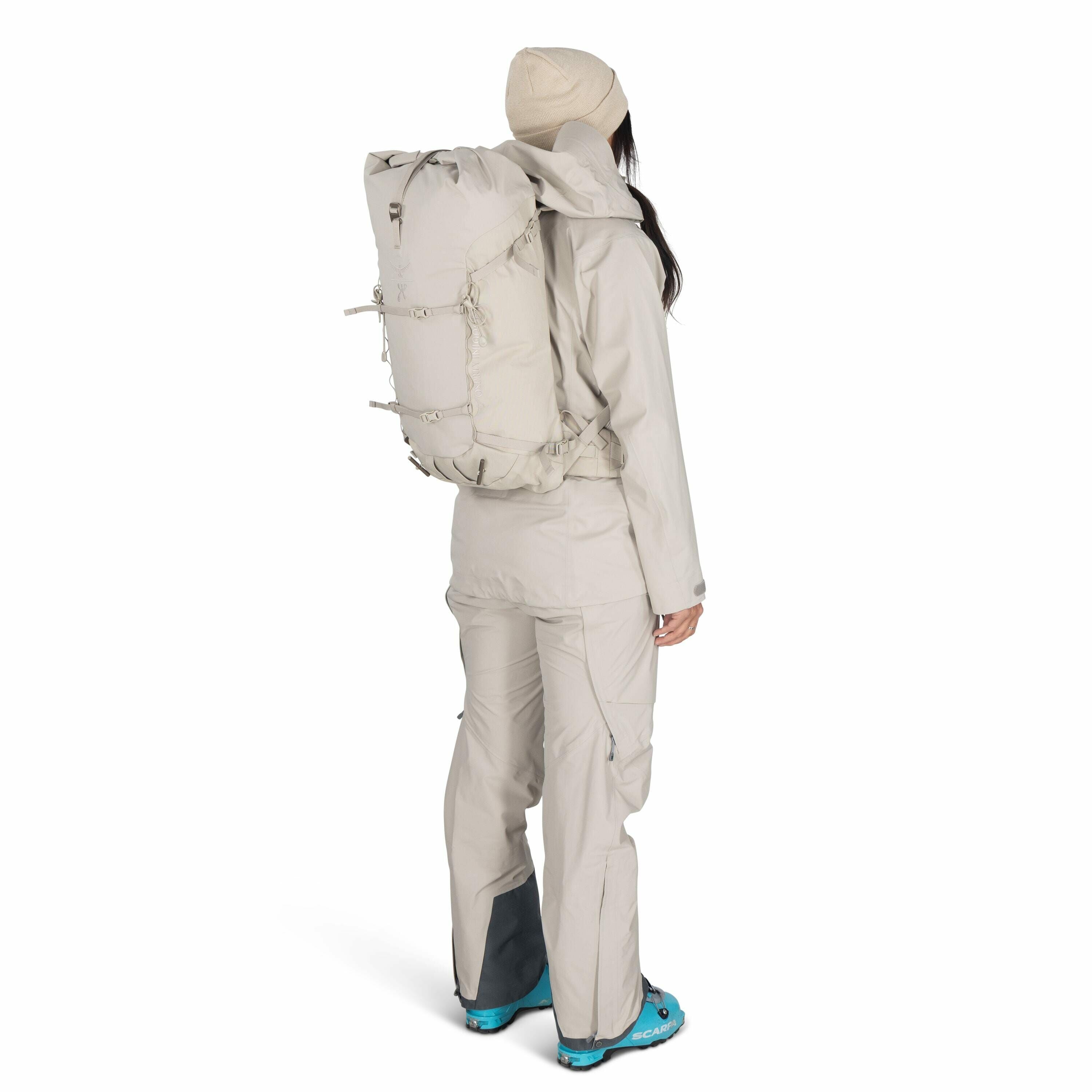 The image features a person dressed in light beige outdoor gear, including a jacket, pants, and a beanie, standing with their back facing the camera. They are wearing a large, beige backpack, which appears to be designed for hiking or mountaineering. The