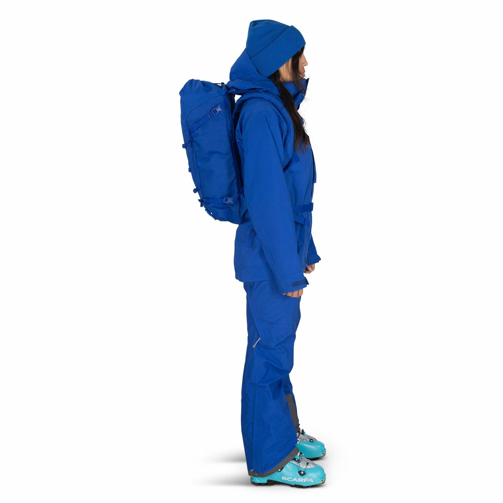 The image features a woman standing in profile, fully dressed in blue outdoor gear. She is wearing a blue jacket, blue pants, and a blue beanie, with a matching blue backpack on her back. Her boots are a light blue color, indicating they may be ski boots