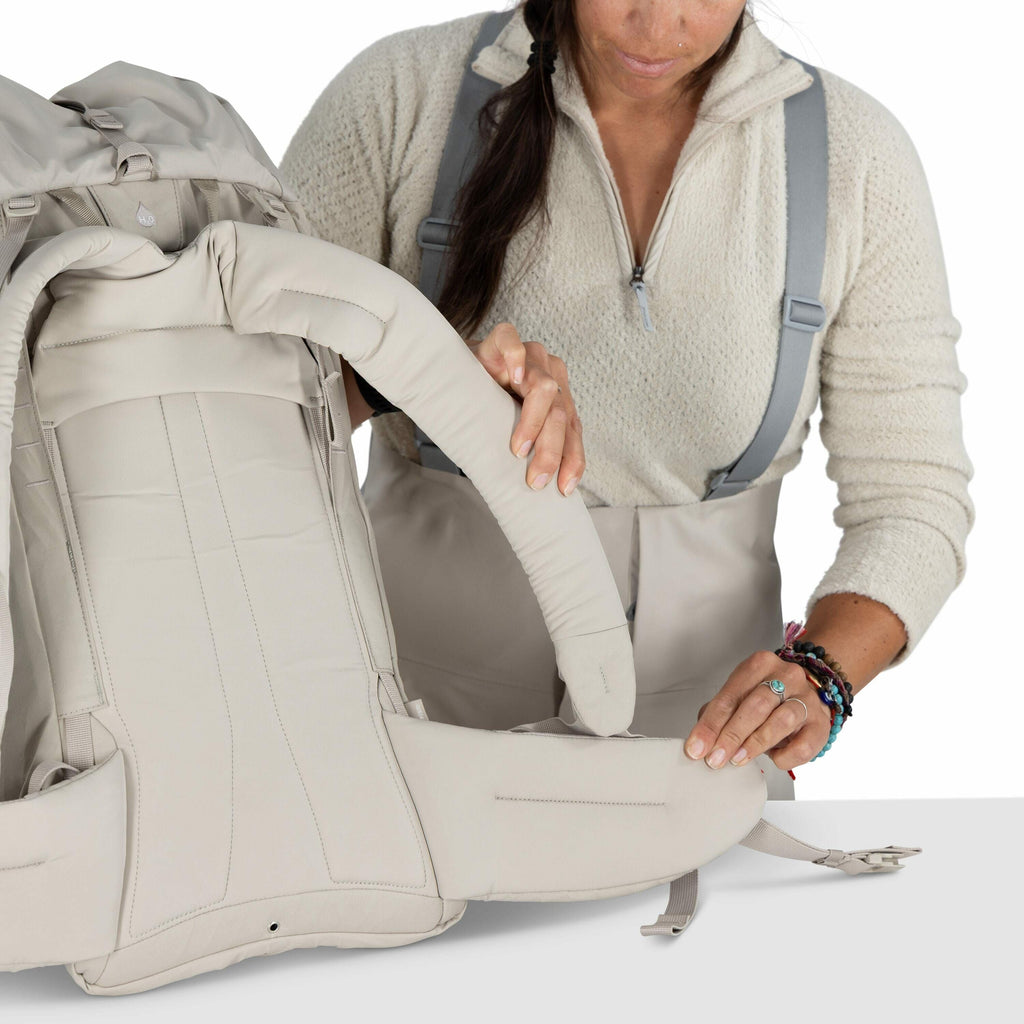 The image shows a woman assembling or adjusting a light beige backpack. She is wearing a textured cream-colored zip-up sweater and has her dark hair partially braided. The backpack appears to be designed for hiking or outdoor use, as it has padded straps