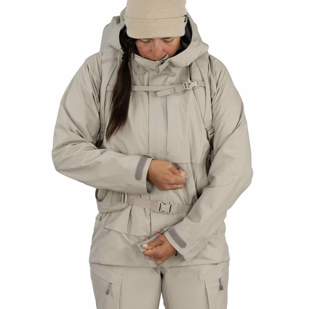 The image shows a person dressed in a light beige outdoor ensemble, consisting of a jacket and pants. The jacket has a high collar and a hood, and features multiple straps and pockets, suggesting it is designed for hiking or outdoor activities. The person