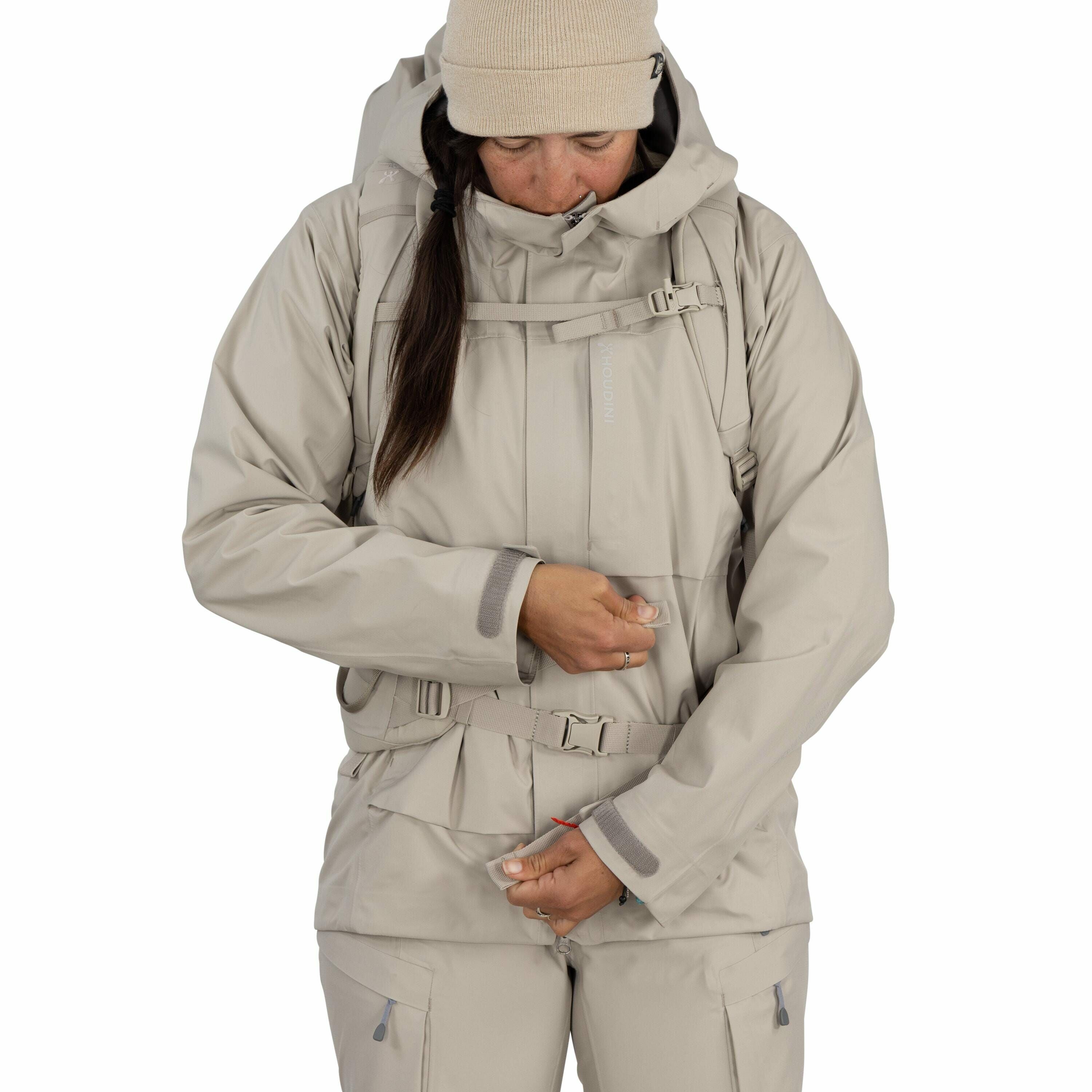 The image shows a person dressed in a light beige outdoor ensemble, consisting of a jacket and pants. The jacket has a high collar and a hood, and features multiple straps and pockets, suggesting it is designed for hiking or outdoor activities. The person