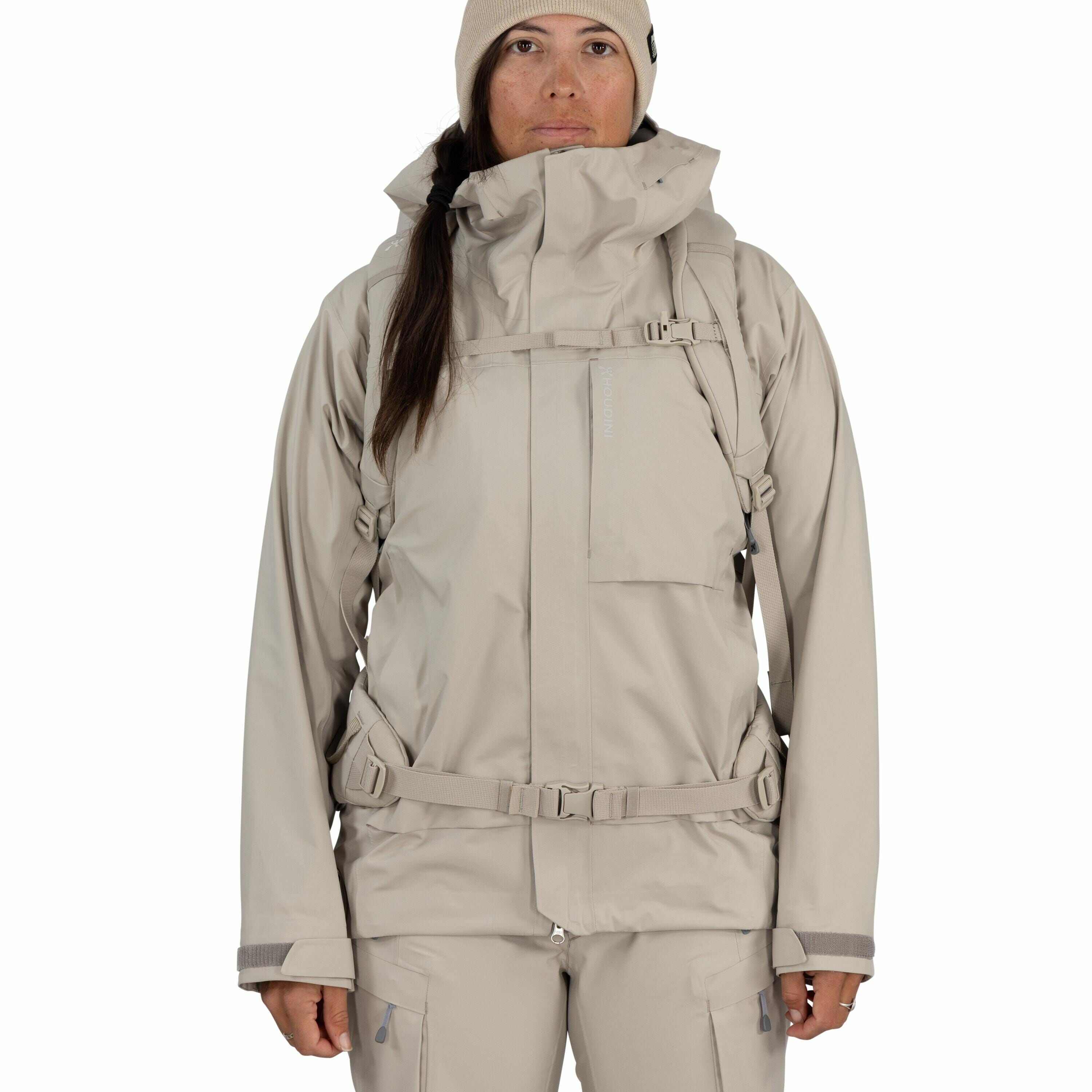 The image depicts a person wearing a light beige, technical outerwear ensemble suitable for cold or wet weather conditions. The outfit consists of a hooded jacket and matching pants, made from what appears to be water-resistant material. The jacket featur