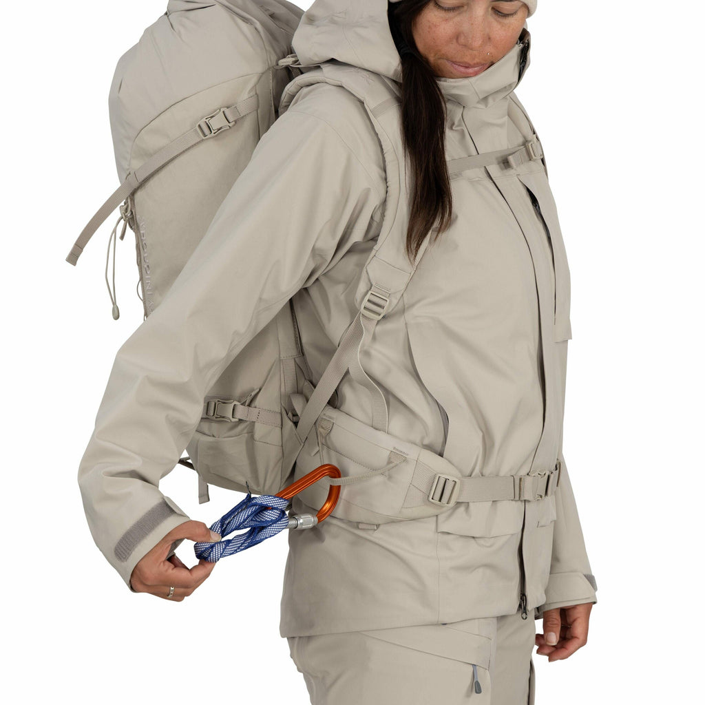 The image shows a person dressed in light grey or beige outdoor gear, consisting of a hooded jacket and matching pants. They are carrying a large backpack on their back, which is equipped with multiple straps and appears to be designed for hiking or climb