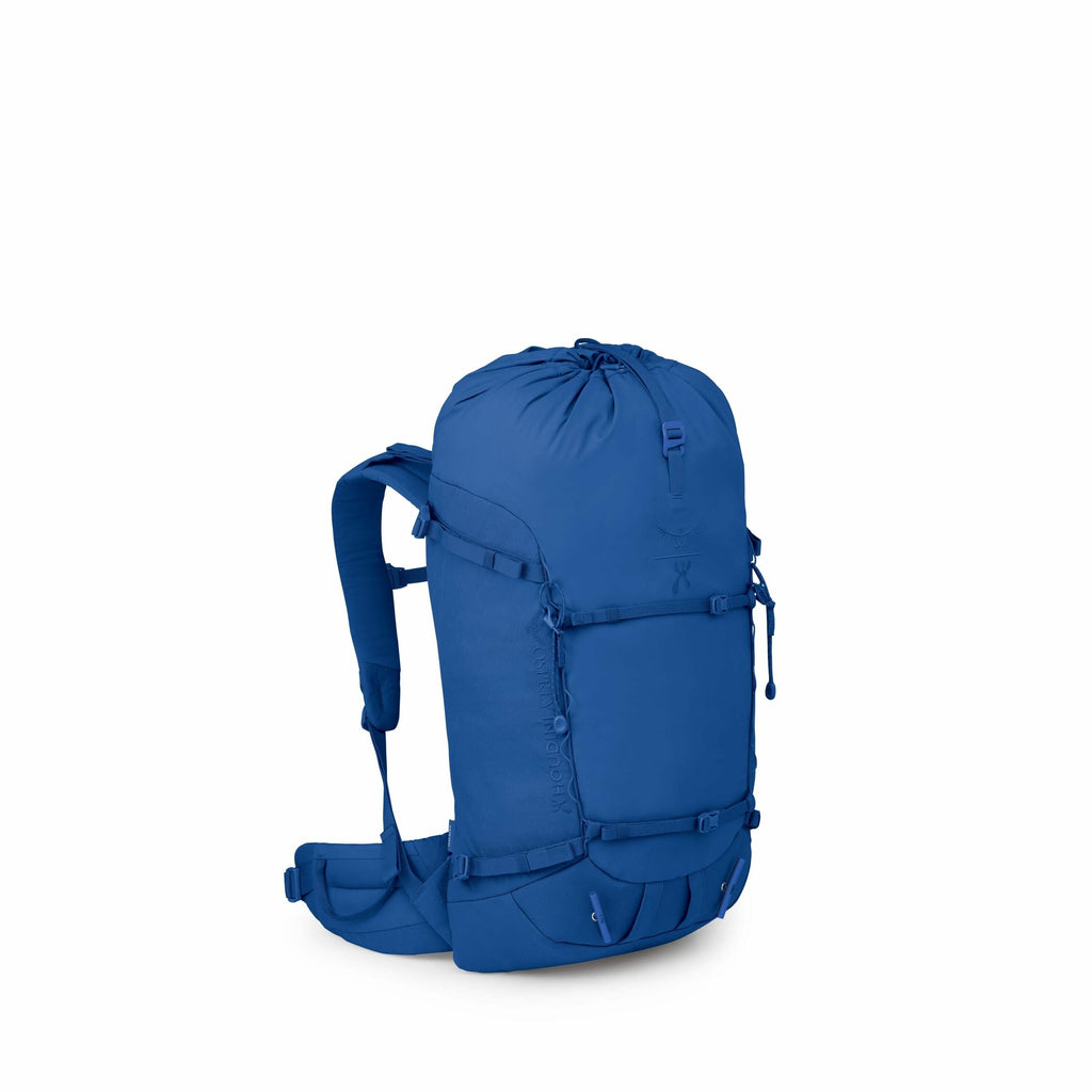The image shows a blue Osprey Mutant 38 climbing backpack designed for outdoor activities like mountaineering or hiking. The backpack has a streamlined and minimalist design with various attachment points and straps for securing gear. It features a top-lo