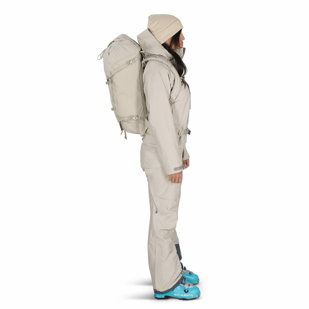 The image shows a woman standing in profile view dressed in outdoor gear. She is wearing a beige hooded jacket and matching pants, complemented by a beige backpack strapped securely on her back. On her head is a beige beanie, coordinating with her outfit.