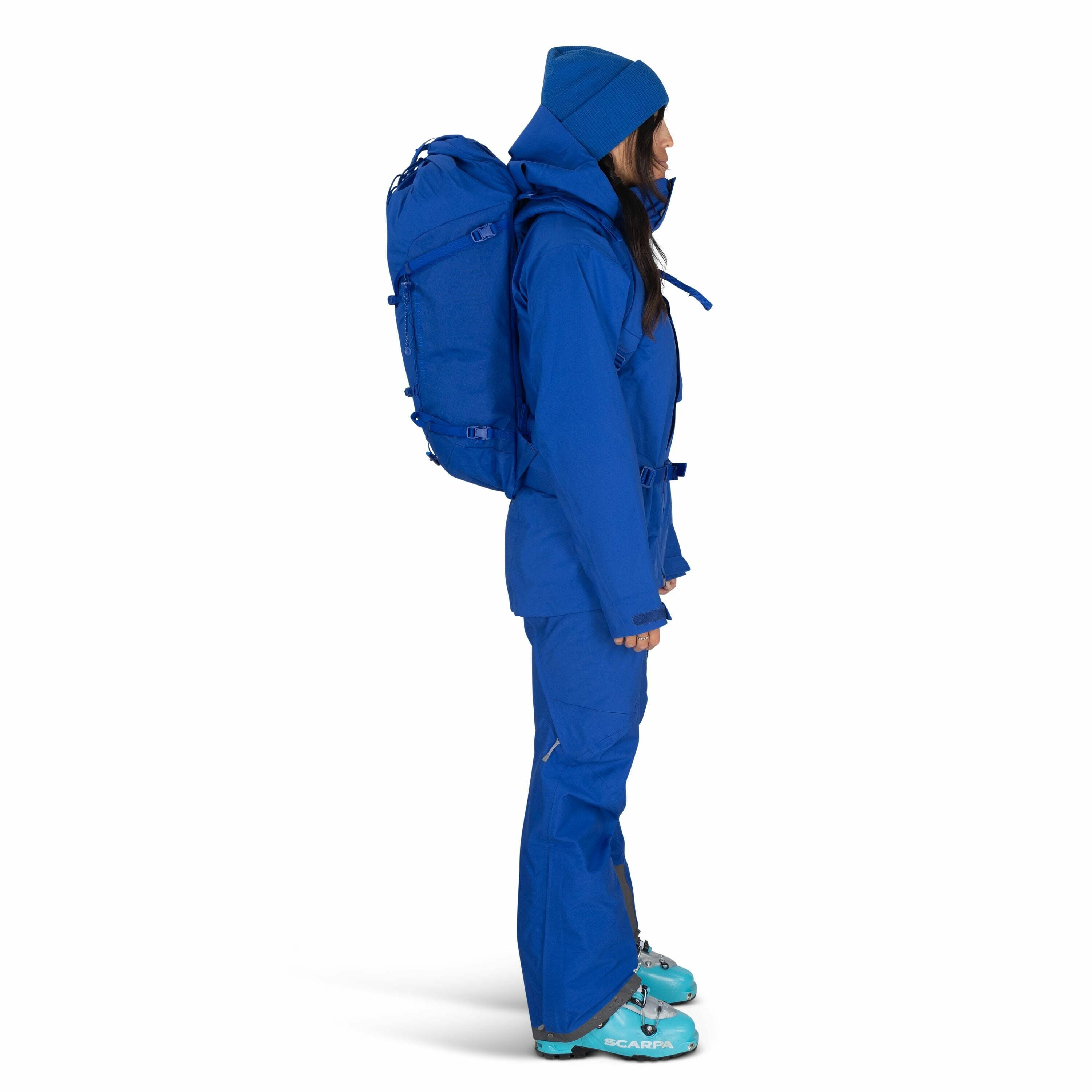 The image shows a person standing in profile view, dressed in a complete set of bright blue outdoor gear. They are wearing a hooded jacket and matching pants, both designed for winter or mountain environments, which suggests that they are prepared for col