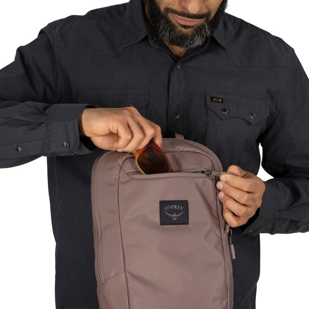 The image shows a person holding open a backpack, displaying its contents. The individual is wearing a dark, button-up shirt with flap pockets on the chest, and they have a wristwatch on their left wrist. Inside the backpack, an electronic device, likely