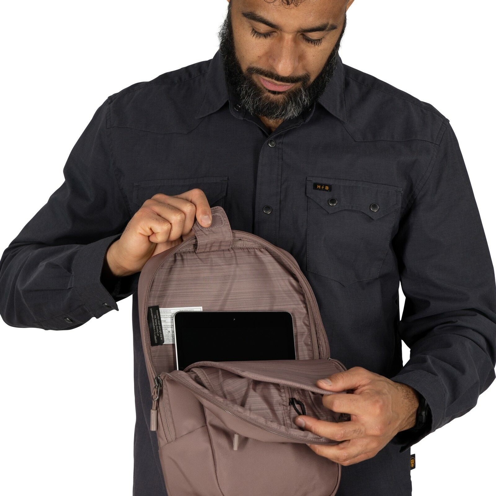 The image depicts a person wearing a dark, long-sleeved button-up shirt, holding a backpack that is partially unzipped. Inside the backpack, the individual is placing or retrieving a pair of red-handled pliers, which are attached to a black keychain or st