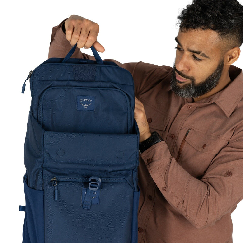 The image shows a person holding a blue Osprey backpack by its top handle, against a plain white background. The backpack features a sleek and modern design with multiple compartments, including a zippered front pocket and a buckle closure for additional