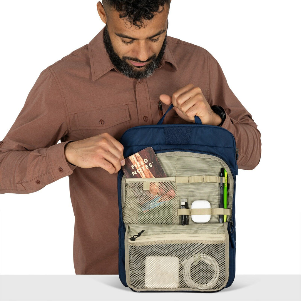 The image features a person wearing a brown button-up shirt holding an organized blue and beige bag by its top handle. The bag has a front section with several mesh and solid pockets, each containing various items like a "Field Notes" notebook, a pair of