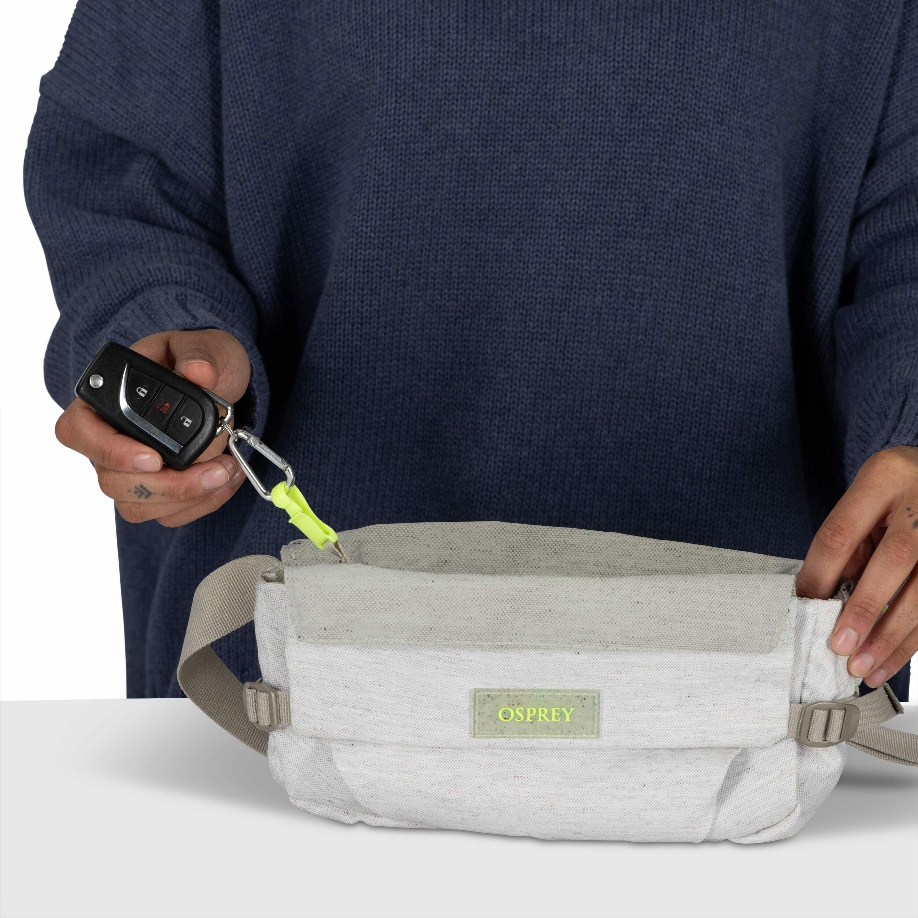 The image depicts a person wearing a dark blue sweater, holding a car key with a keychain in one hand and opening a pouch with the other. The pouch is a light gray color with a beige top, featuring a strap for carrying around the waist. The pouch bears a