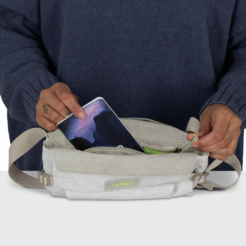 The image shows a person dressed in a dark blue sweater, holding a light-colored Osprey waist pack. The person's left hand is seen placing or retrieving a smartphone, which displays a scenic wallpaper, from the main compartment of the bag. The right hand
