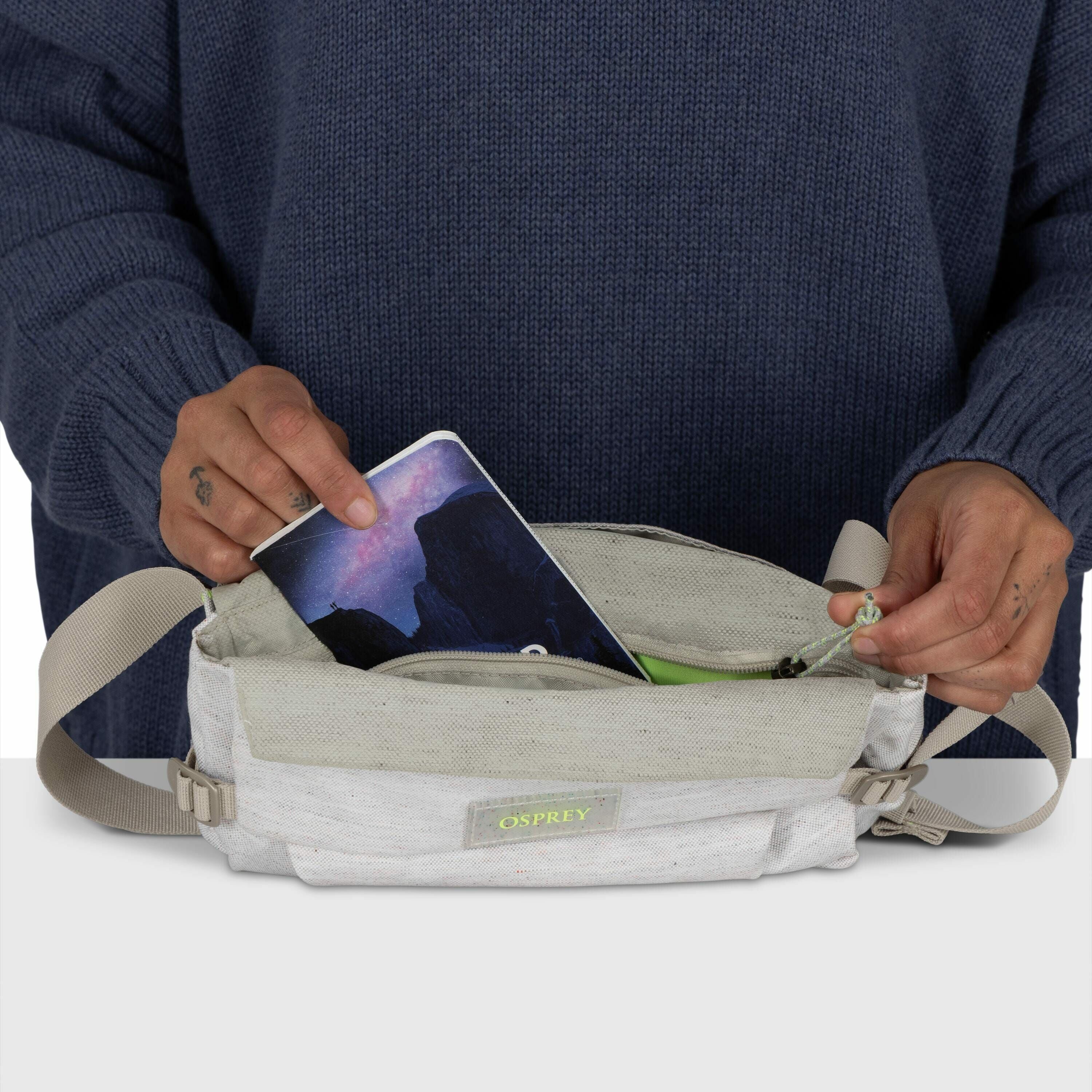 The image shows a person dressed in a dark blue sweater, holding a light-colored Osprey waist pack. The person's left hand is seen placing or retrieving a smartphone, which displays a scenic wallpaper, from the main compartment of the bag. The right hand