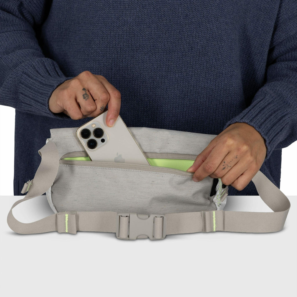 The image features a person wearing a blue sweater, holding a light gray fabric waist bag with one hand while using the other hand to insert or retrieve a smartphone from its zippered compartment. The smartphone is a modern model, likely an iPhone, with t
