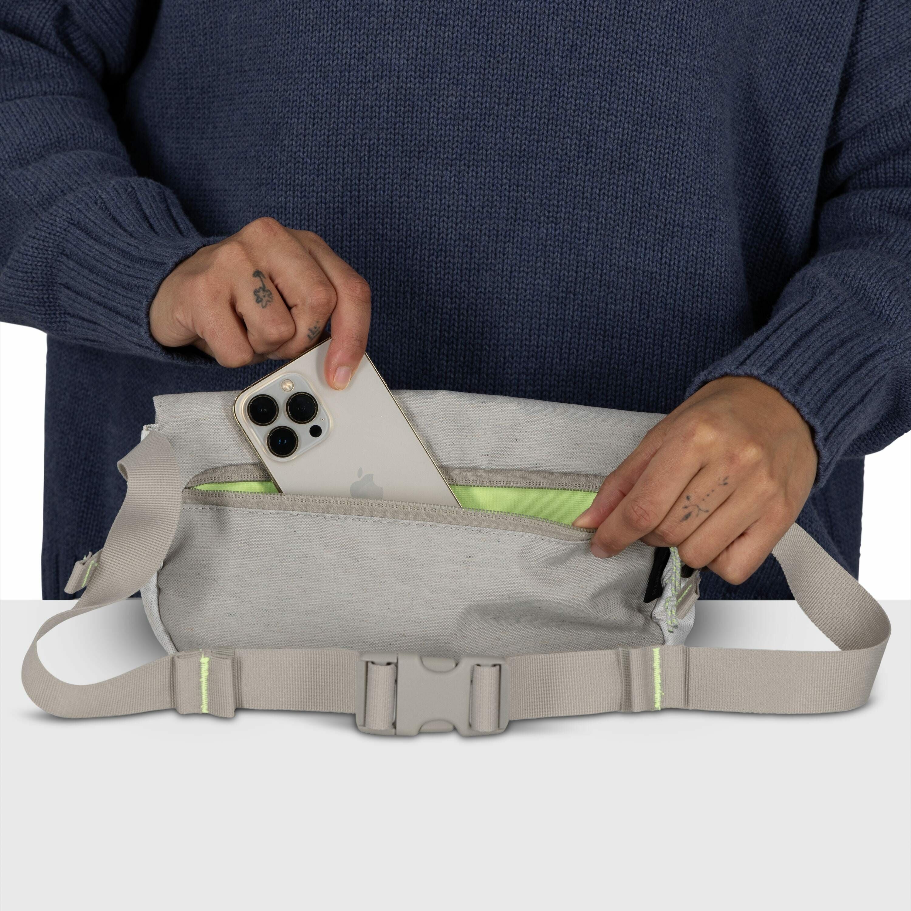 The image features a person wearing a blue sweater, holding a light gray fabric waist bag with one hand while using the other hand to insert or retrieve a smartphone from its zippered compartment. The smartphone is a modern model, likely an iPhone, with t