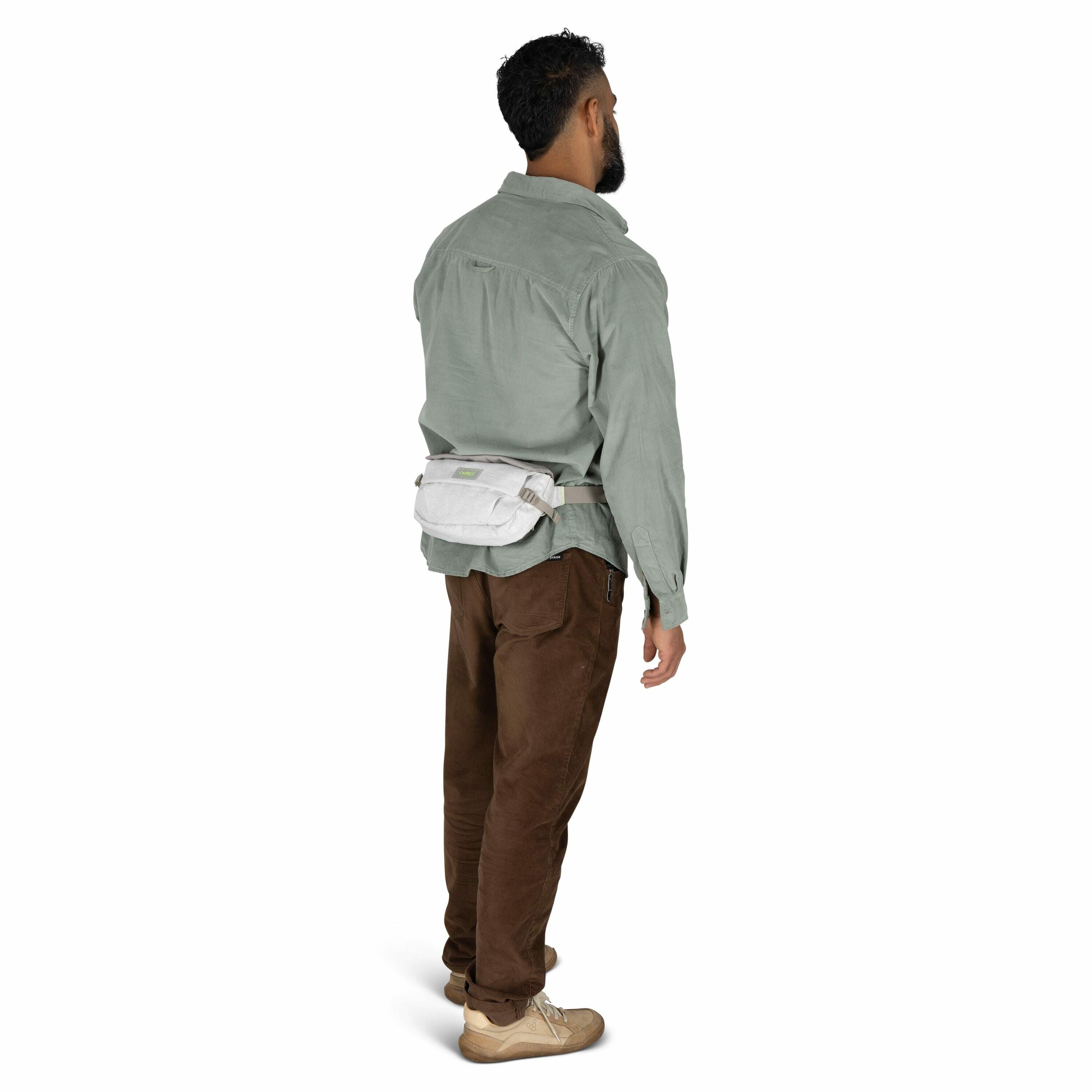 The image shows a man standing and facing away from the camera, wearing casual, outdoorsy clothing. He has a thick beard and is dressed in a light green button-up shirt and brown pants with a slight sheen. On his waist, he has a light gray or off-white wa