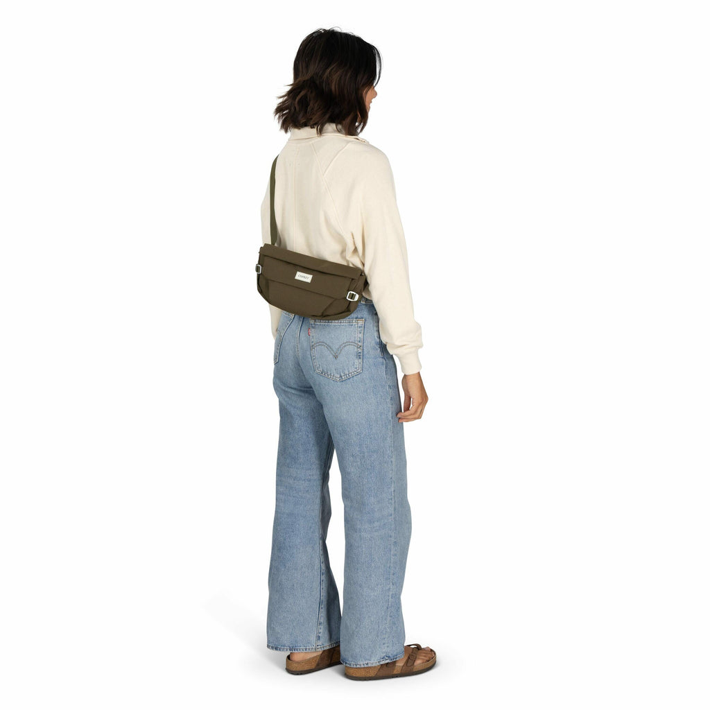 The image depicts a woman standing with her back turned to the camera. She is wearing a cream-colored long-sleeve top and high-waisted light blue denim jeans with flared legs. A khaki green crossbody bag is slung across her back, featuring a label from th