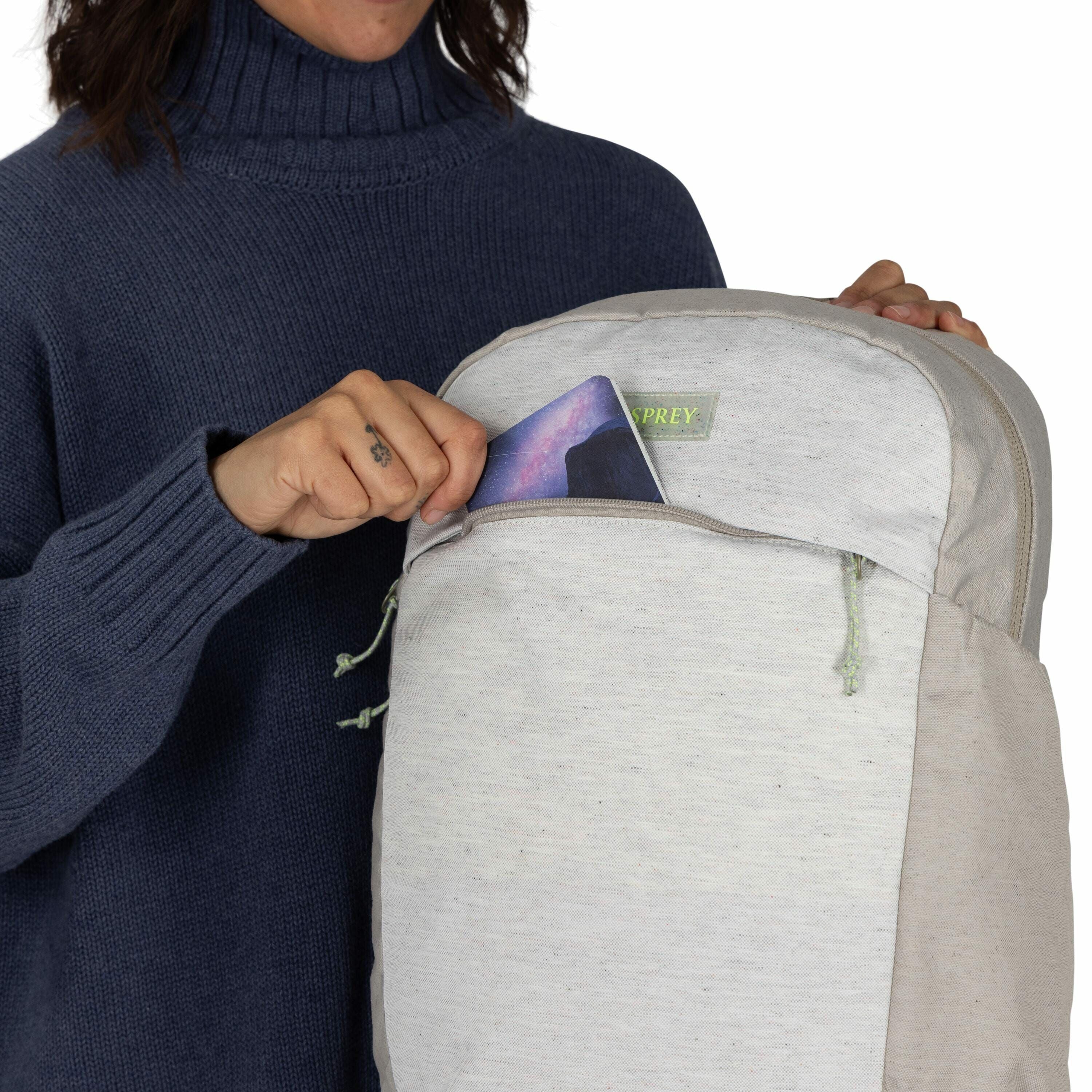 The image shows a person placing or retrieving a tablet or similar electronic device from the front pocket of a backpack. The backpack is primarily light gray, with a subtle, green "OSPREY" logo on the front. The person is wearing a dark blue, ribbed turt