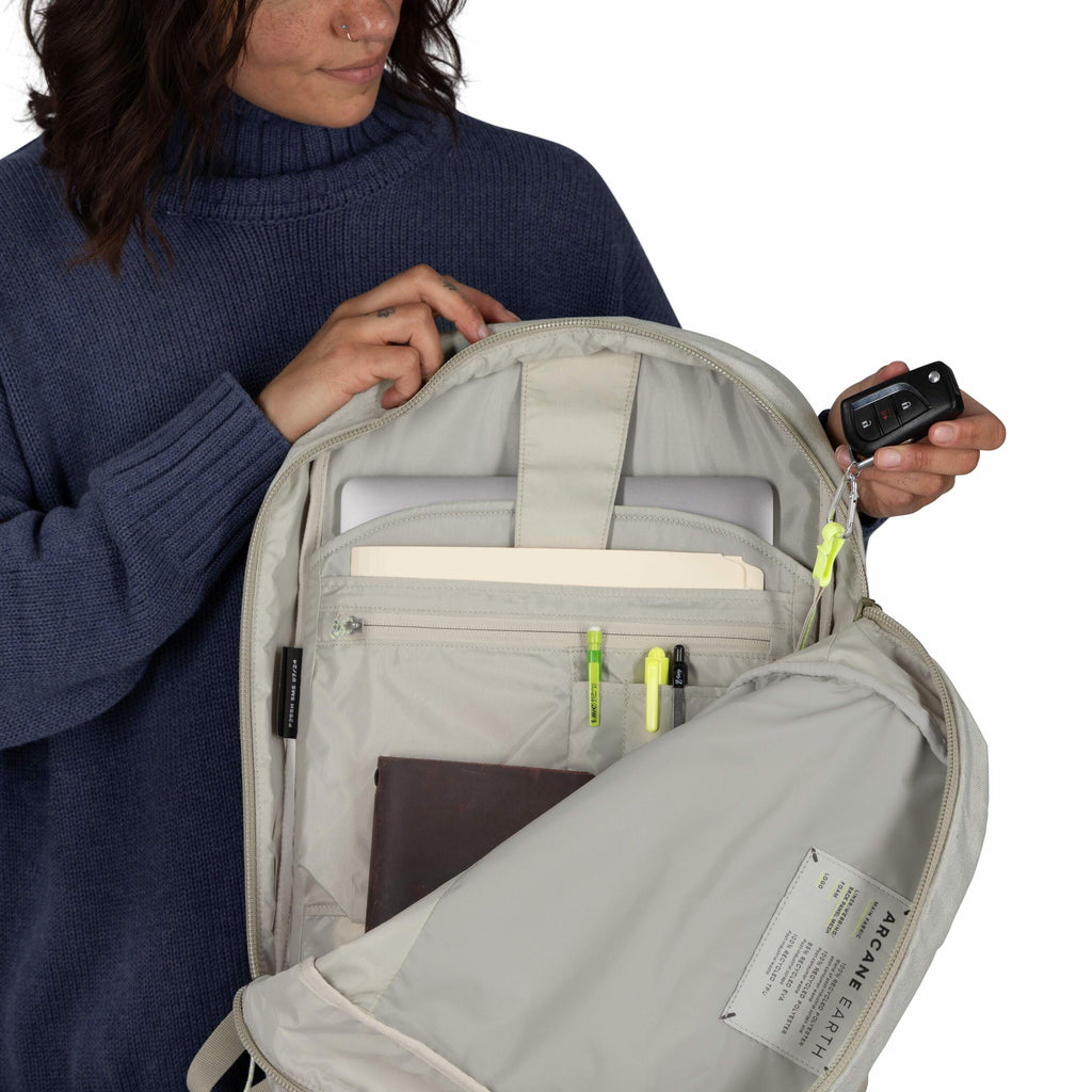 The image shows a woman holding open a light-colored backpack to reveal its contents. Inside the backpack, there is a laptop in a dedicated sleeve, folders, and several pens attached to an organizer panel. She is also holding a set of car keys, highlighti
