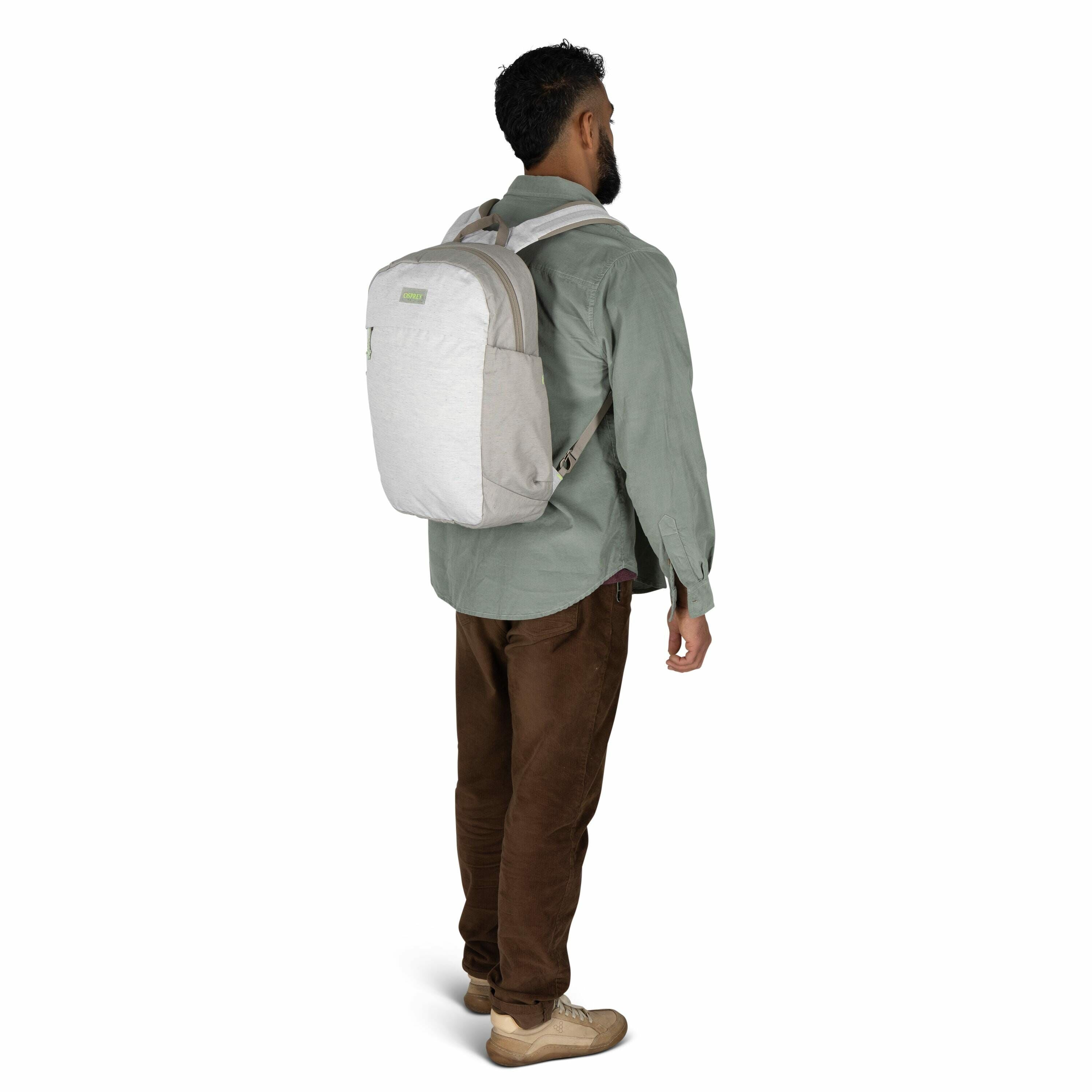 The image features a man standing and facing away from the camera, showcasing a backpack. He is wearing a light green button-up shirt and dark brown pants, with tan-colored shoes. The backpack he is carrying is light grey with beige accents and appears to