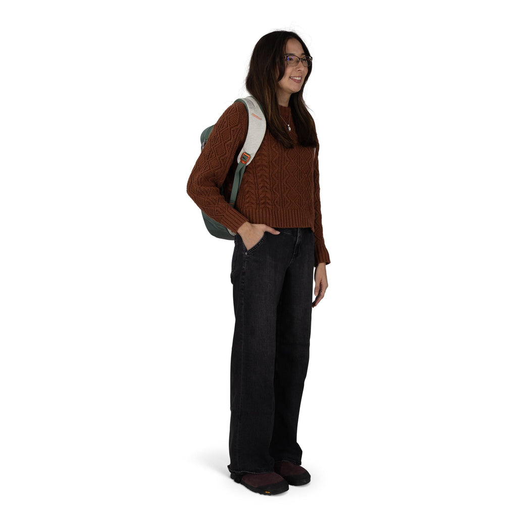 A person stands in a casual pose with one hand in a pocket, wearing a textured brown cable-knit sweater and loose black jeans. They have dark hair and wear sunglasses, and a light-gray-strapped green backpack over one shoulder. The outfit is finished with