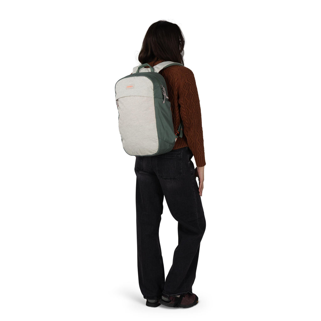 A person is shown from the back wearing a two-tone backpack in muted green and light gray. They are dressed in a brown cable-knit sweater, loose black jeans, and dark shoes. The backpack has a front zip pocket and a small logo patch near the top. The back