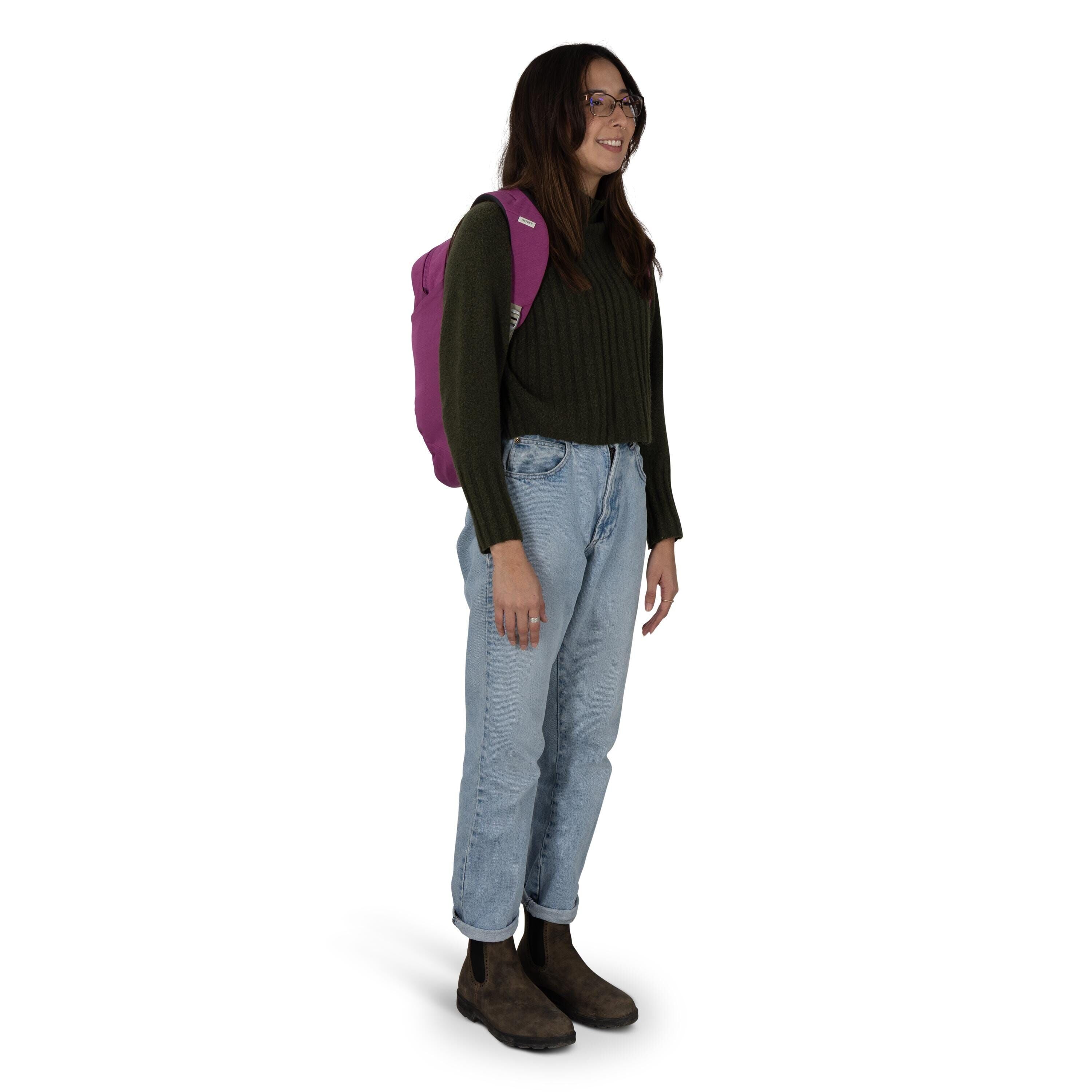A person stands against a white background wearing casual, outdoor-inspired attire. They have on a dark green ribbed sweater, light blue cuffed jeans, and brown Chelsea-style boots. A magenta backpack is slung over one shoulder. The stance is relaxed, wit