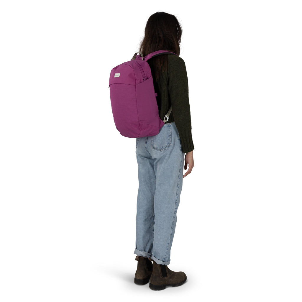 A person is standing in a three-quarter back view, wearing a magenta backpack. They have on a dark green sweater, light blue cuffed jeans, and brown ankle boots. The backpack has a simple, streamlined shape with a small rectangular logo patch on the upper