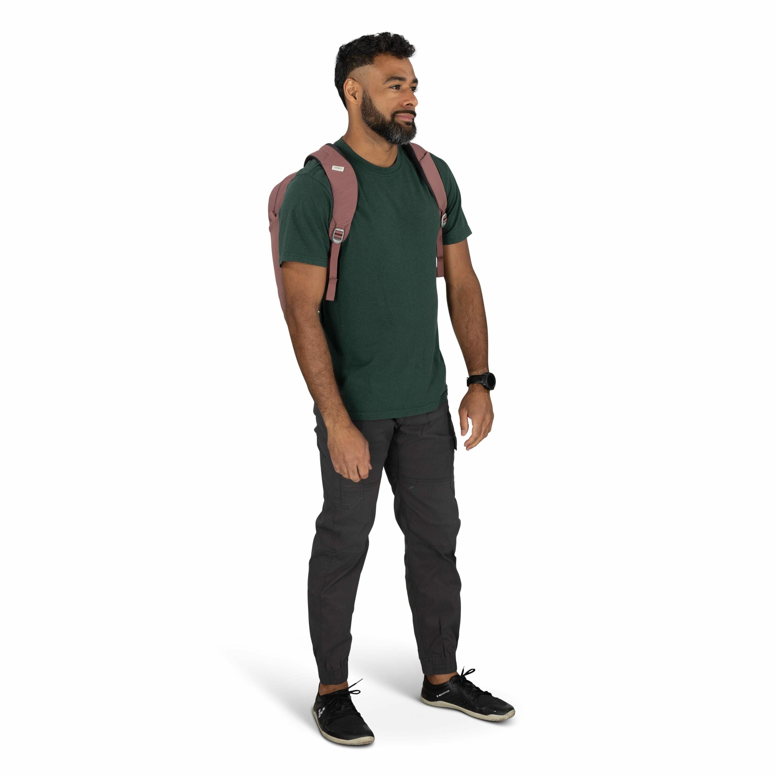 The image shows a man standing with a casual and relaxed posture. He is wearing a forest green t-shirt paired with dark gray cargo pants and black sneakers, giving off a comfortable and laid-back look. The man is carrying a dusty pink backpack with visibl