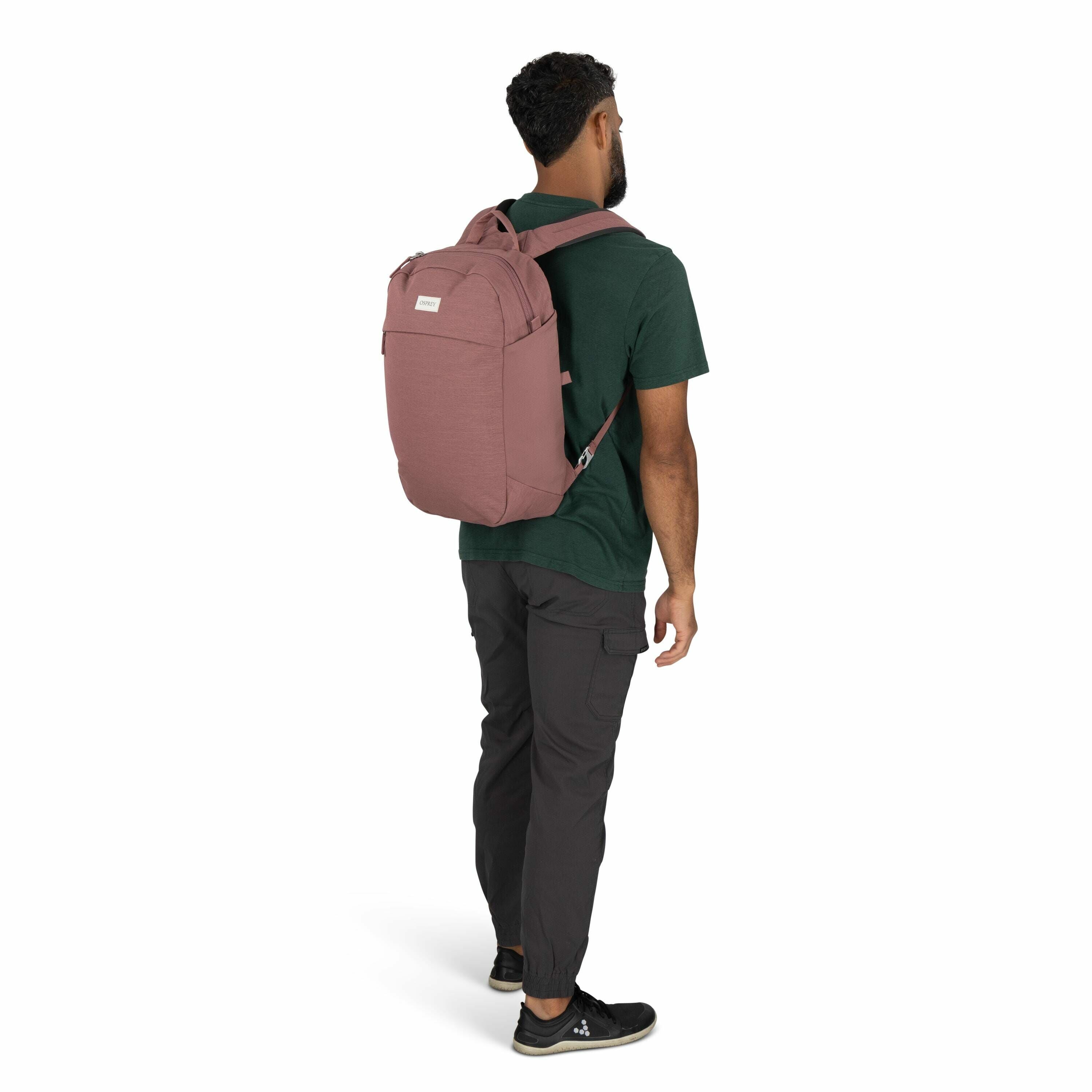 The image depicts a man standing with his back towards the camera, wearing a pink backpack. He has short black curly hair and a beard, and is dressed casually in a dark green t-shirt and dark gray pants with cargo pockets. The backpack is made of fabric,