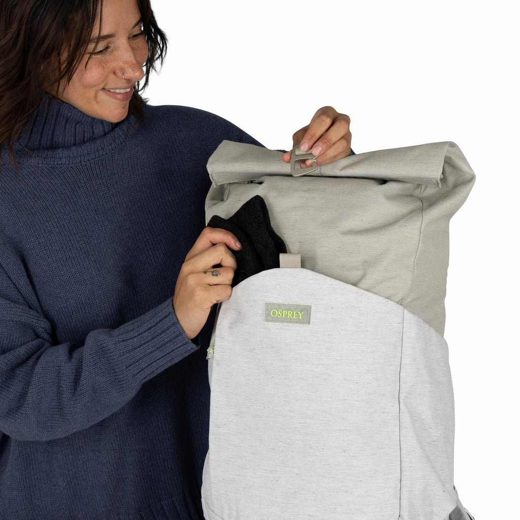 The image shows a person holding a backpack, which is designed with a two-tone color scheme of light gray and a slightly darker gray. The person is wearing a blue, long-sleeved, knit sweater and appears to be placing a small black pouch or item into the b