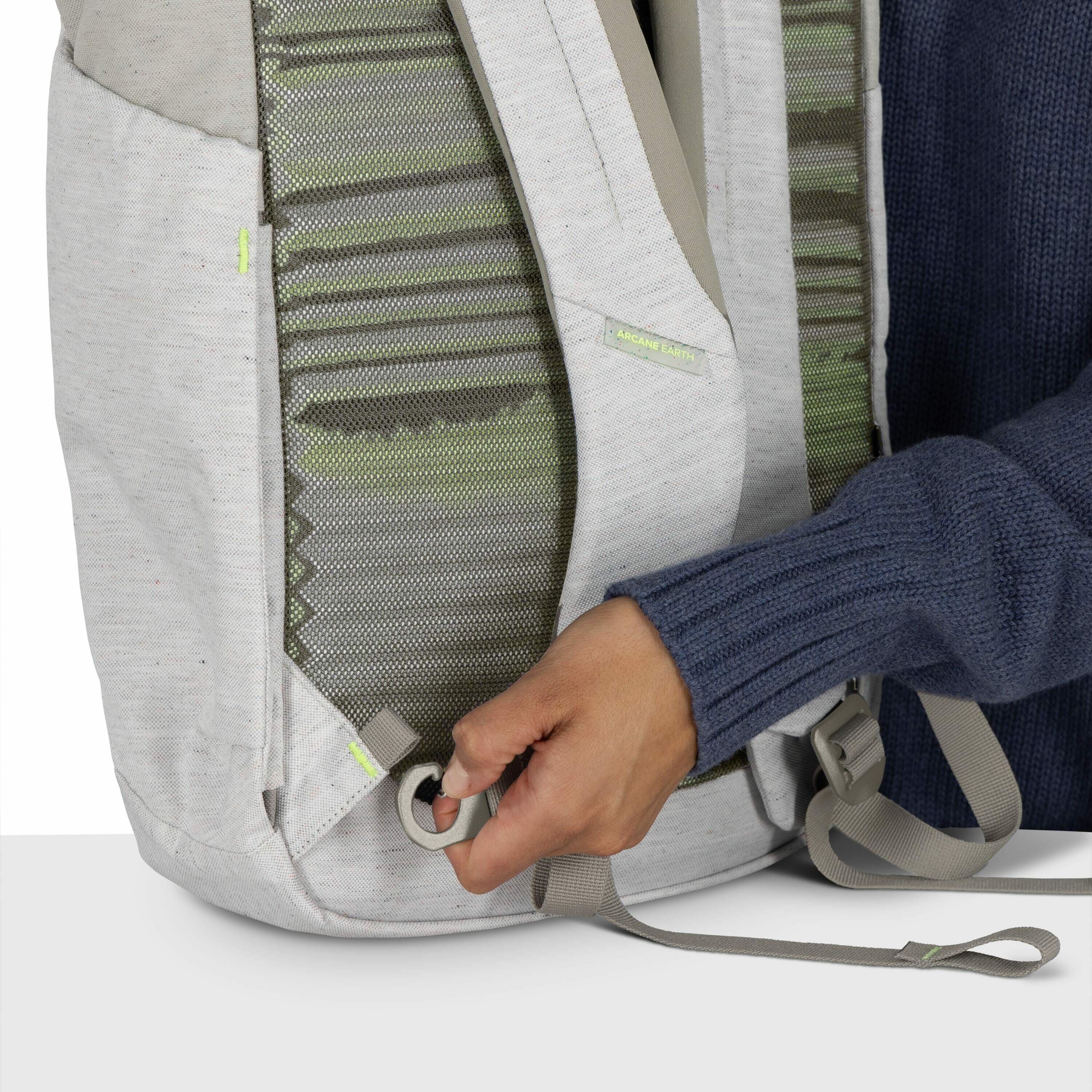The image shows a close-up of a person adjusting or pulling a strap on a light gray backpack. The backpack features green patterned fabric visible between the padded sections of the back panel. The person is wearing a dark blue knitted sweater, and their