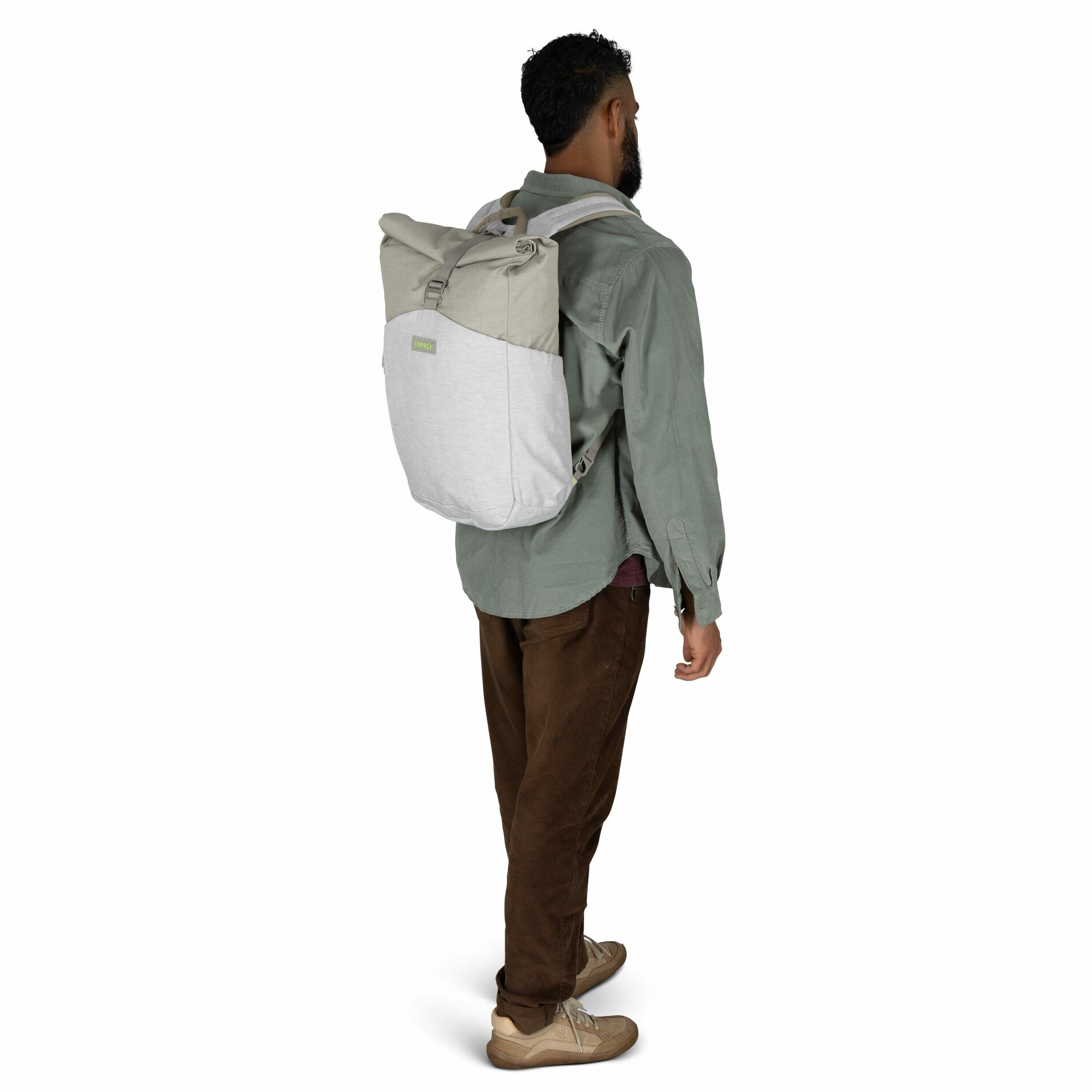 The image features a man standing sideways, showcasing a backpack worn on his back. He has short curly hair and is dressed in casual clothing, including a green long-sleeved button-up shirt and brown pants. The backpack he is wearing is a two-tone design,