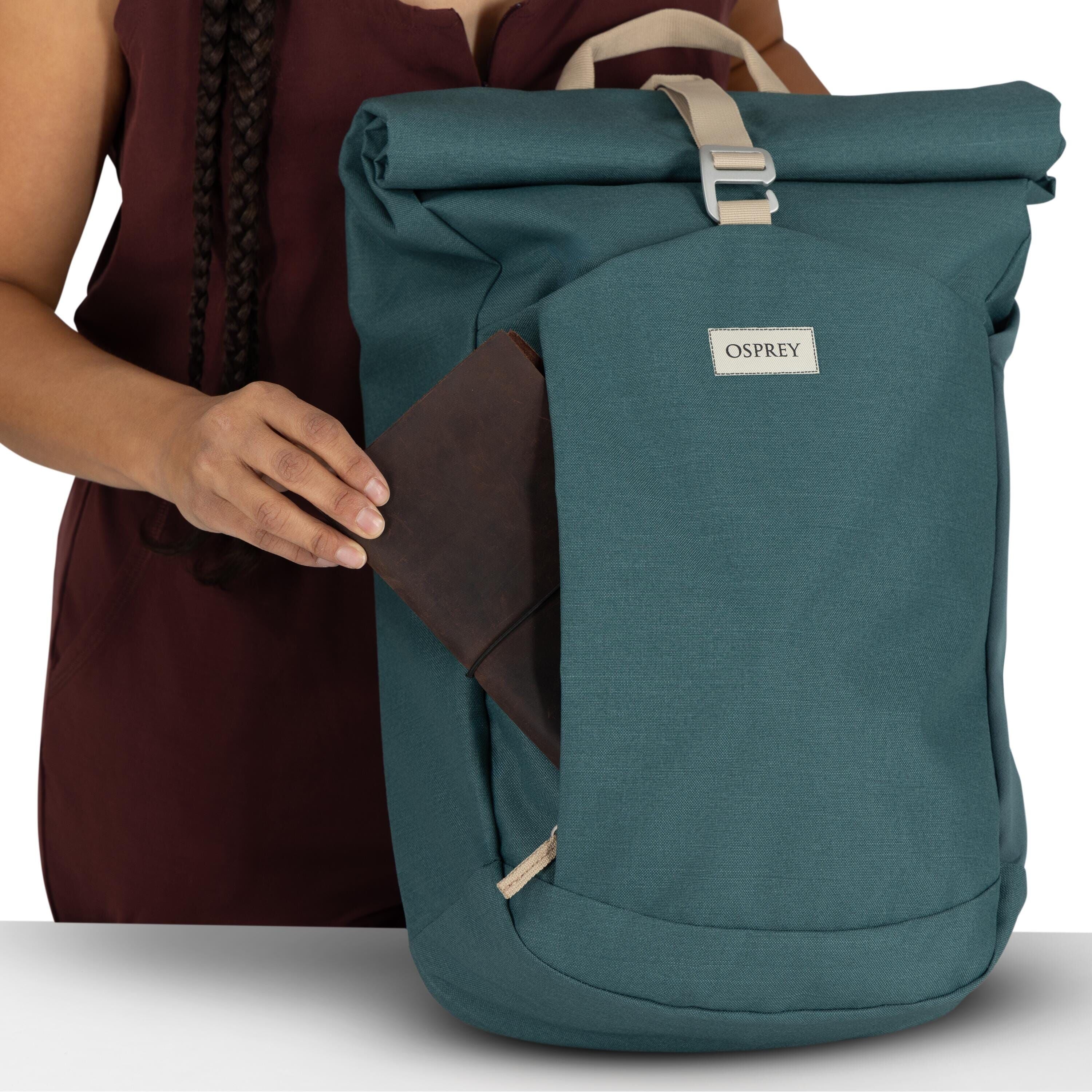 The image features a person wearing a burgundy outfit with braided hair visible, placing or removing a brown leather wallet from a teal Osprey backpack. The backpack has a distinct roll-top design, secured with a beige buckle, and the brand name "OSPREY"