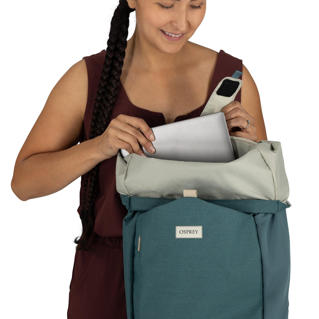 The image features a person dressed in a sleeveless maroon top and is holding a backpack from the brand Osprey. The backpack has a two-tone design with a blue exterior and a light gray top flap. The individual is placing or removing a sleek gray laptop or