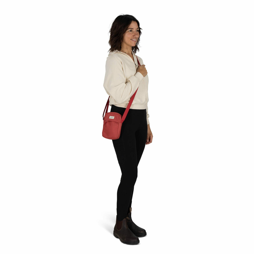 The image features a woman standing against a plain white background. She is wearing a light-colored sweatshirt and black leggings, paired with dark ankle boots. Draped across her body is a small red crossbody bag, which she holds with one hand. Her hair