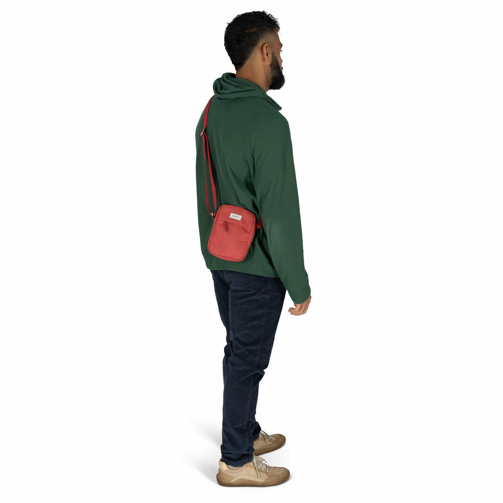 The image features a man standing in a casual pose, turned slightly to his left with his back partially facing the camera. He is wearing a dark green hoodie, dark blue jeans, and beige sneakers. A small red crossbody bag is slung over his right shoulder,