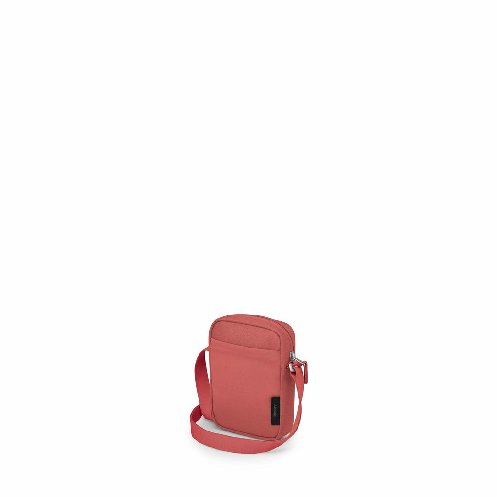 The image displays a small, red crossbody bag with a minimalist design. The bag has a front pocket with a zipper closure and a main compartment that appears to also have a zip closure. It features an adjustable shoulder strap for convenient carrying. The