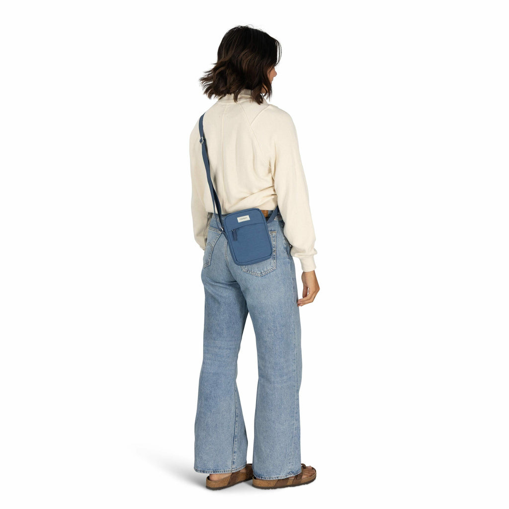 The image features a woman standing with her back facing the camera, dressed in a casual outfit. She is wearing a cream-colored long-sleeve sweater and light blue flared jeans. On her feet are brown slip-on shoes. The woman is carrying a small blue crossb