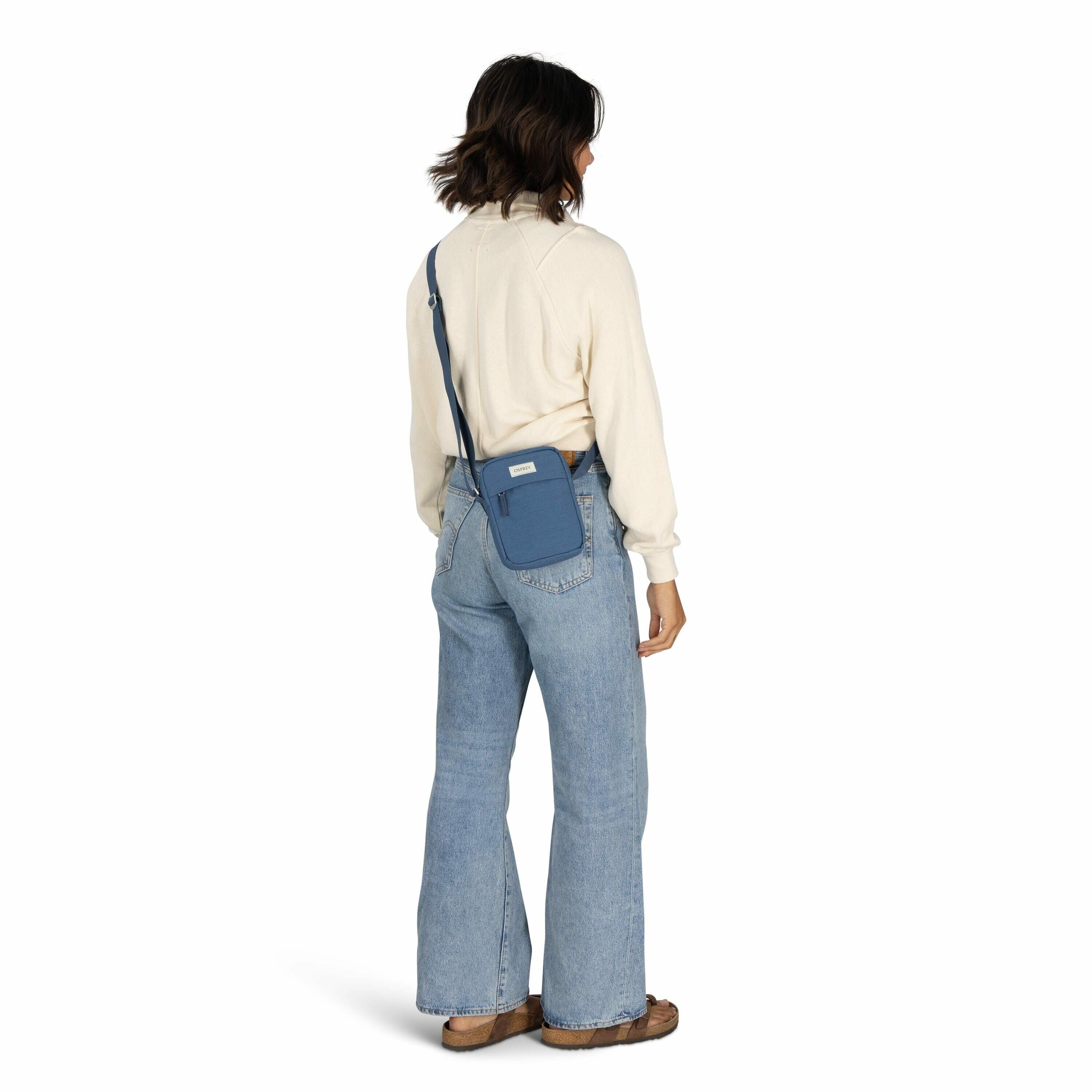 The image features a woman standing with her back facing the camera, dressed in a casual outfit. She is wearing a cream-colored long-sleeve sweater and light blue flared jeans. On her feet are brown slip-on shoes. The woman is carrying a small blue crossb