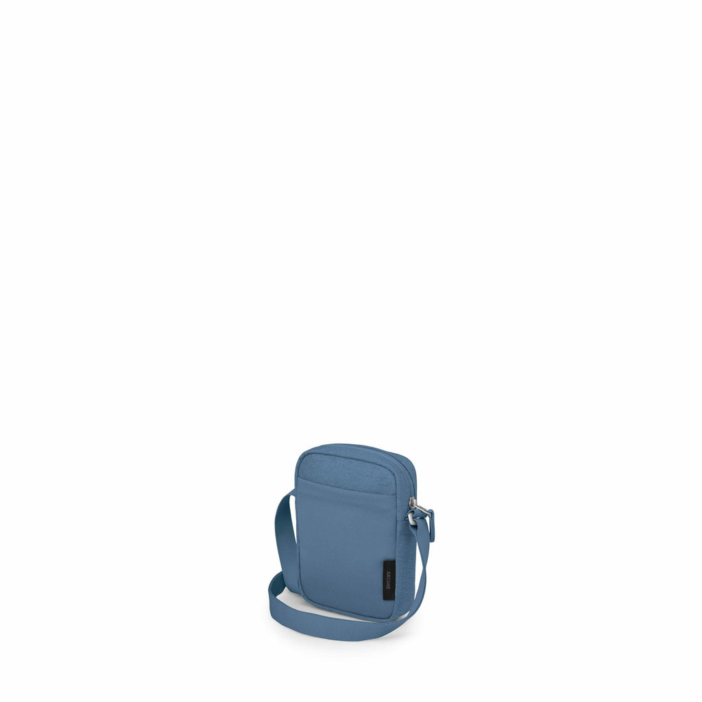 The image displays a compact crossbody bag in a muted blue color. The bag has a simple, modern design with clean lines and a textured fabric exterior. It features a single main compartment with a zip closure, and there's a visible black label on the front