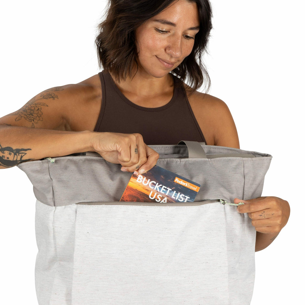 The image features a person wearing a sleeveless brown top and holding a large, light grey bag with darker grey accents. The person's arm is adorned with floral tattoos. They are placing a travel guide titled "Bucket List USA" from Fodor's Travel into the