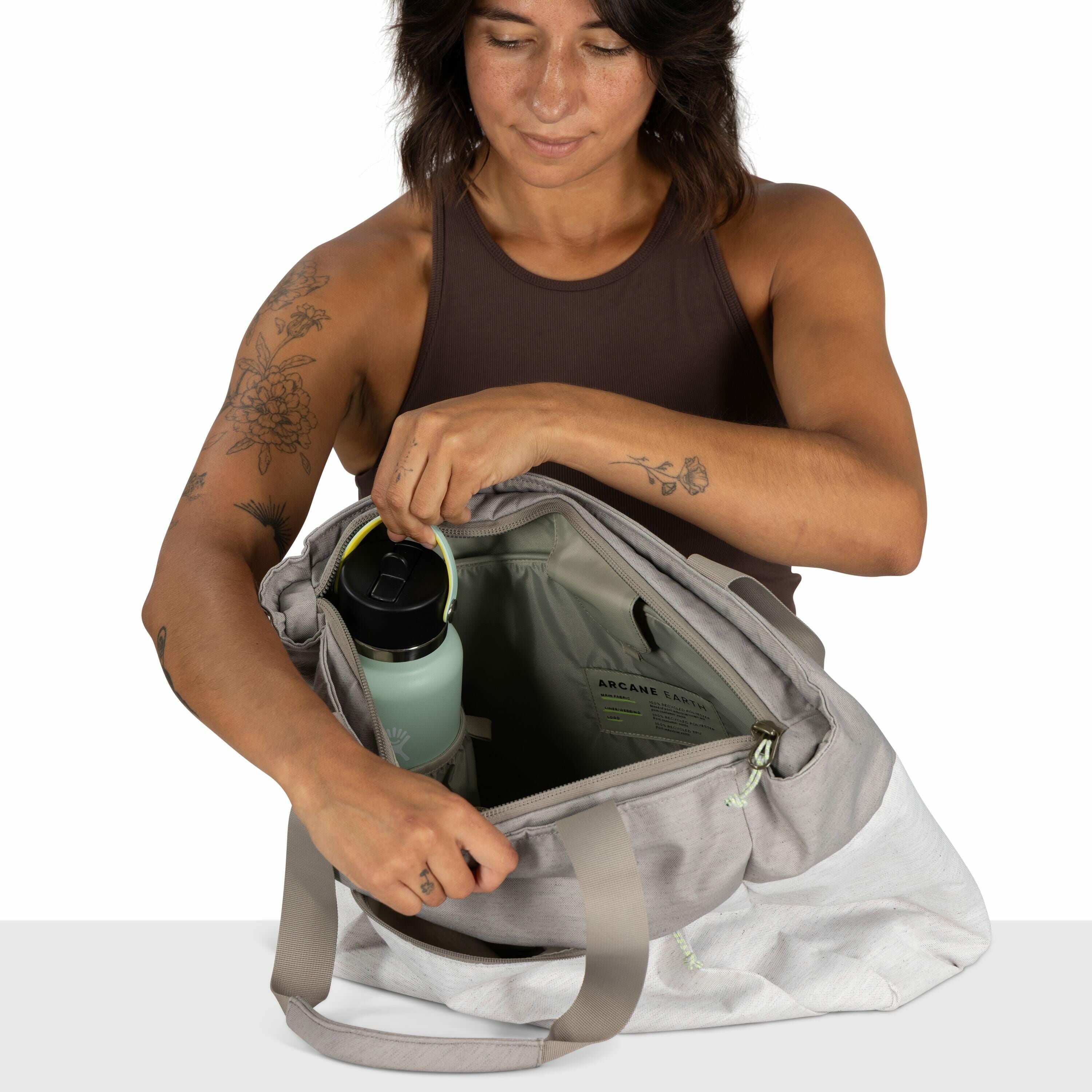 The image shows a person holding a gray and white tote bag, with their other hand grasping a light blue water bottle inside the bag. The individual is wearing a dark sleeveless top, revealing arms adorned with various tattoos, including floral designs and